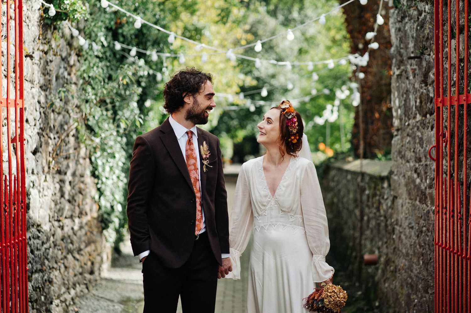 A bride and groom hold hands and smile at each other in a charming outdoor alley adorned with string lights.