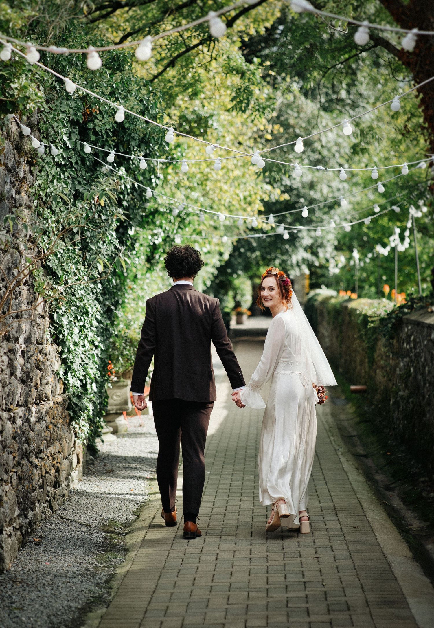 Bride and groom walking hand in hand down a charming ivy-lined pathway with string lights overhead.