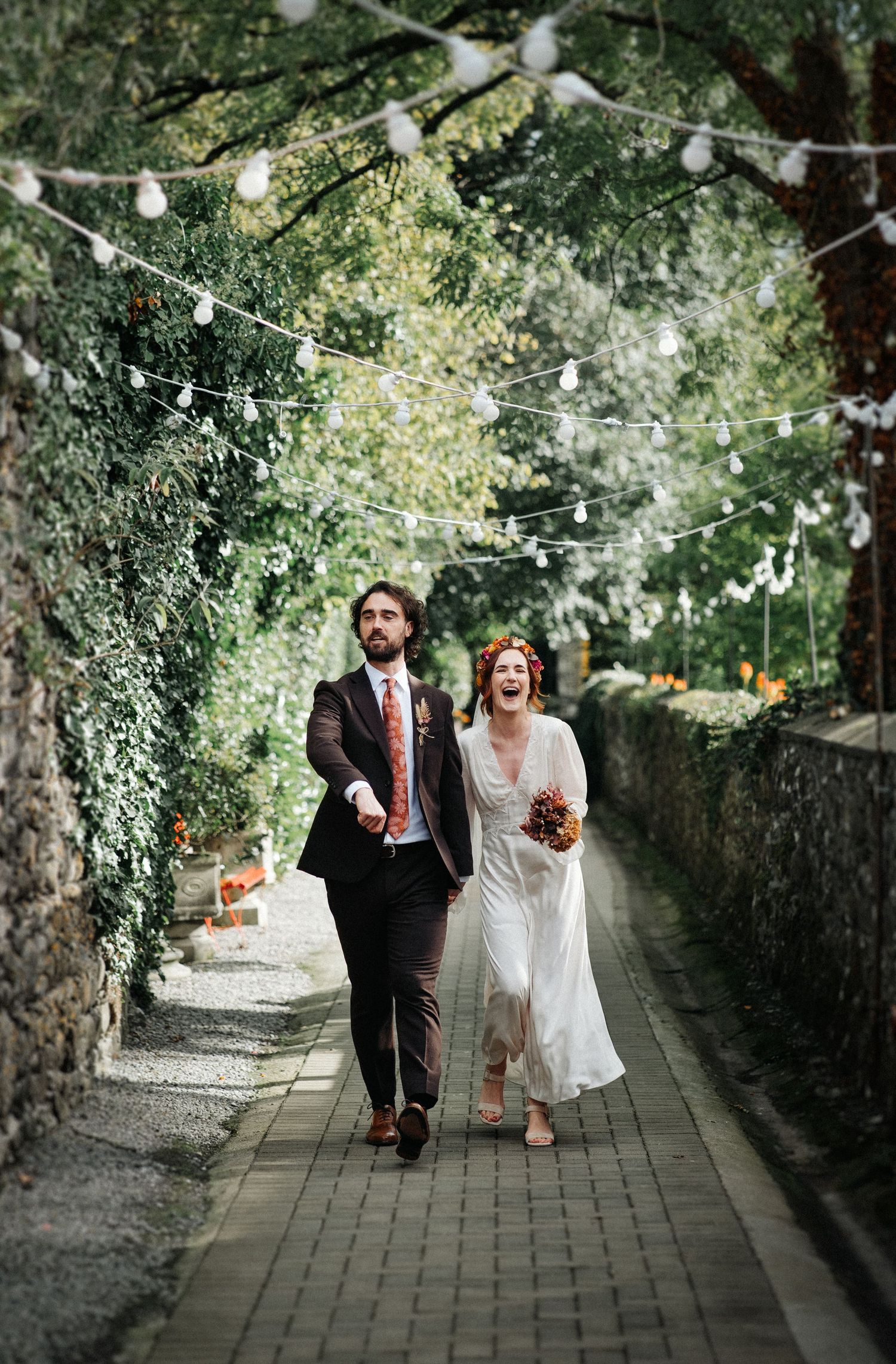 A joyful bride and groom walk hand-in-hand down a charming garden pathway adorned with string lights.