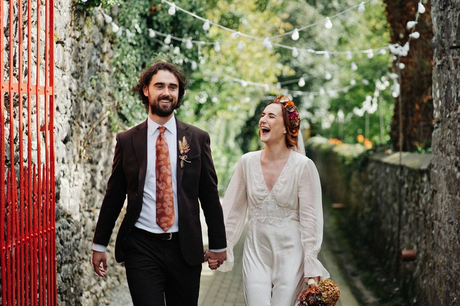Happy couple walks hand-in-hand down a charming alley with string lights, bride in white jumpsuit, groom in dark suit with floral tie.