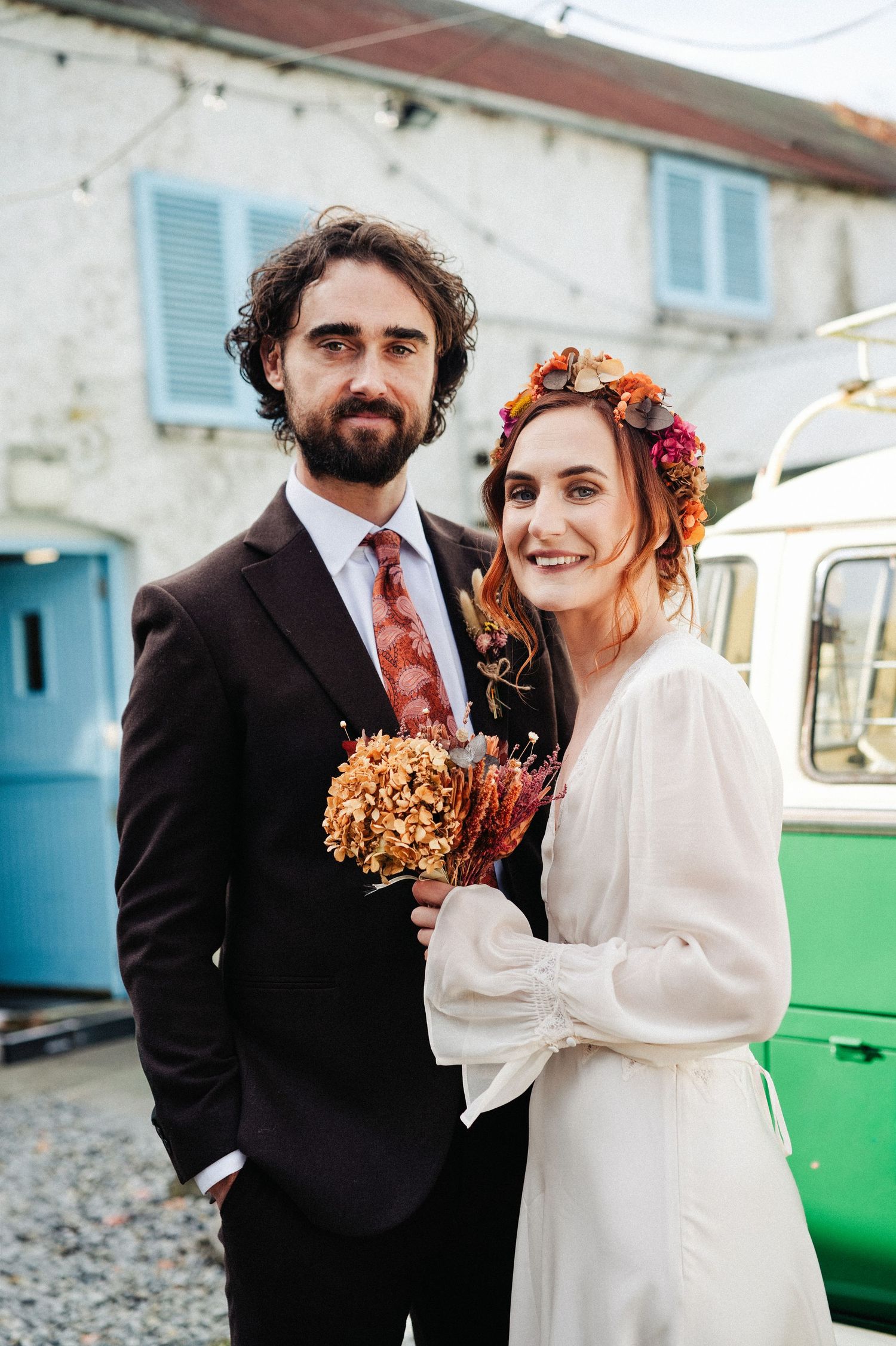 Bride and groom pose outdoors, she wears a floral crown and white dress, he wears a dark suit with floral tie.