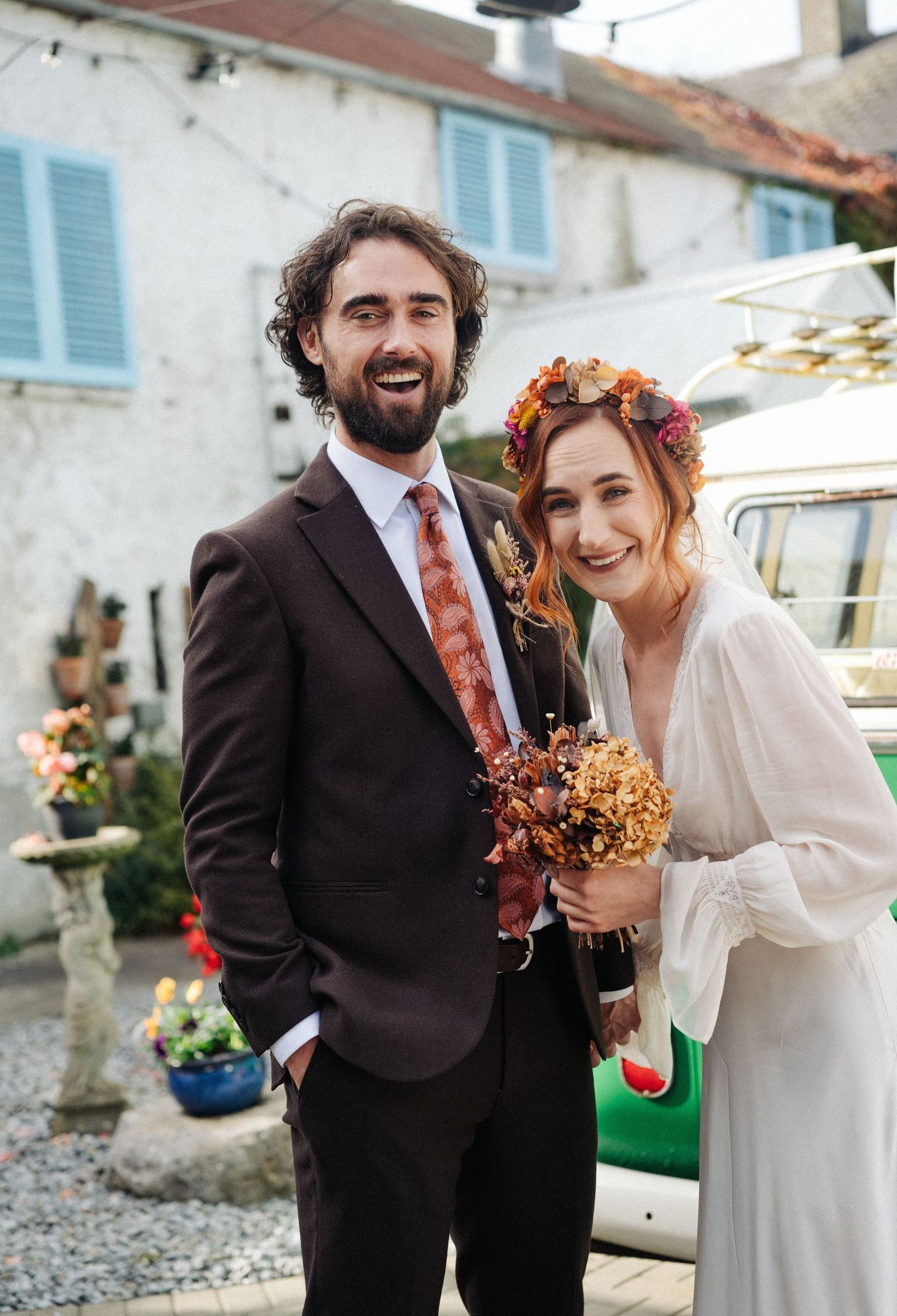 A smiling couple in wedding attire, groom in dark suit with floral tie, bride in white dress with autumn flower crown.