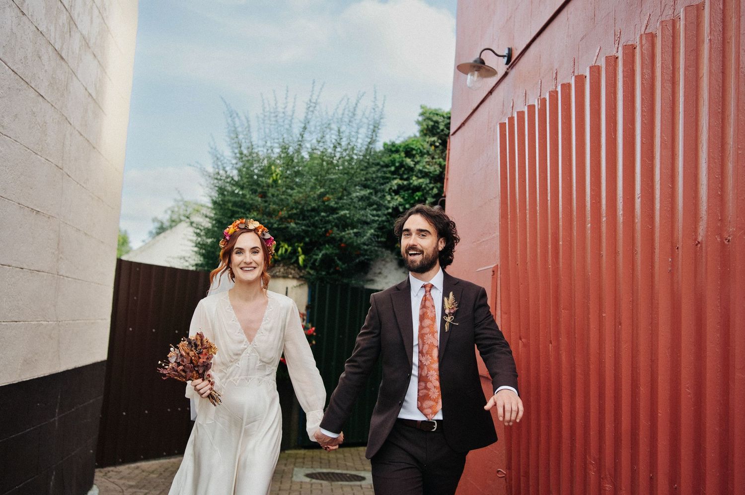 Bride and groom walk hand-in-hand past a red wall, she in white lace gown with floral crown, he in dark suit.
