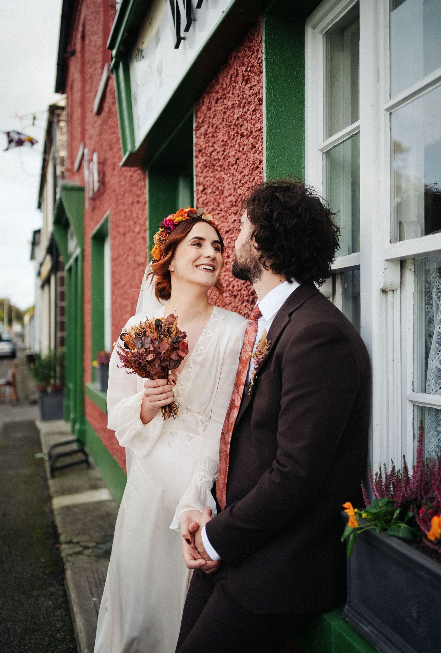 Bride in white dress with autumn flower crown holds bouquet as groom in dark suit whispers to her outside colorful building.