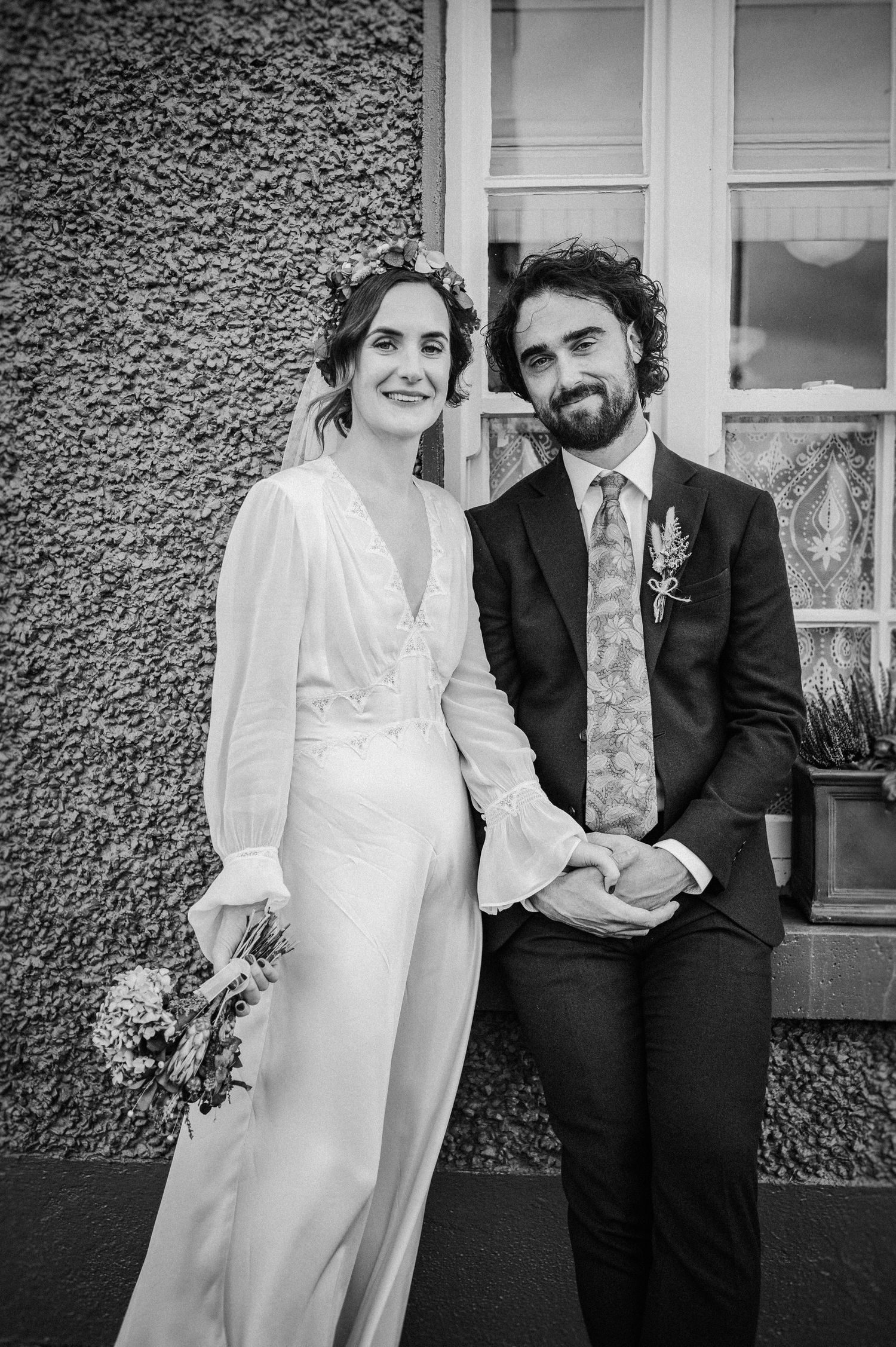 Black and white wedding photo of a smiling bride in white gown with flower crown and groom in dark suit with floral tie.