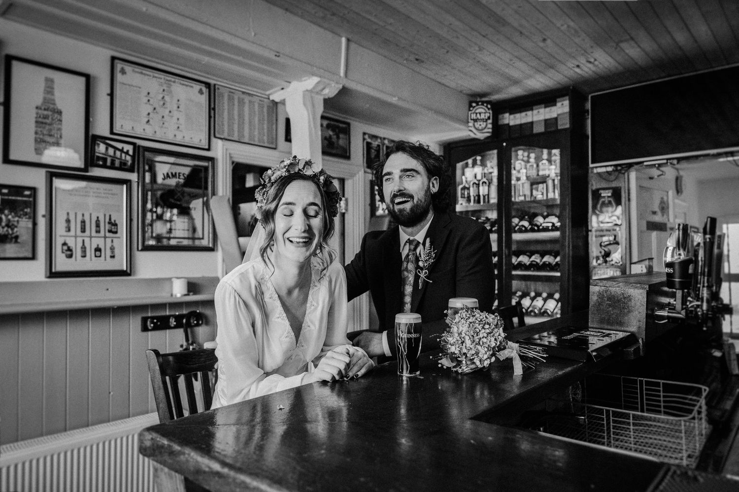 Black and white photo of a smiling couple at a pub bar, woman in bridal attire, man in suit, celebrating together.