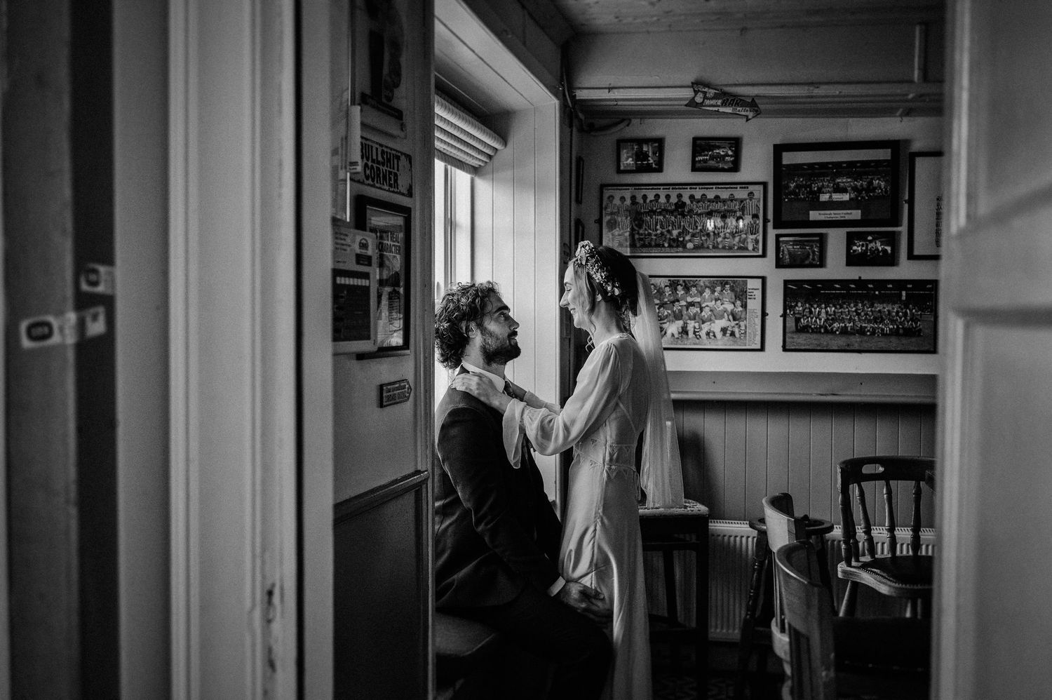 Black and white wedding photo of groom kneeling as bride adjusts his tie in a cozy room with framed photos on walls.