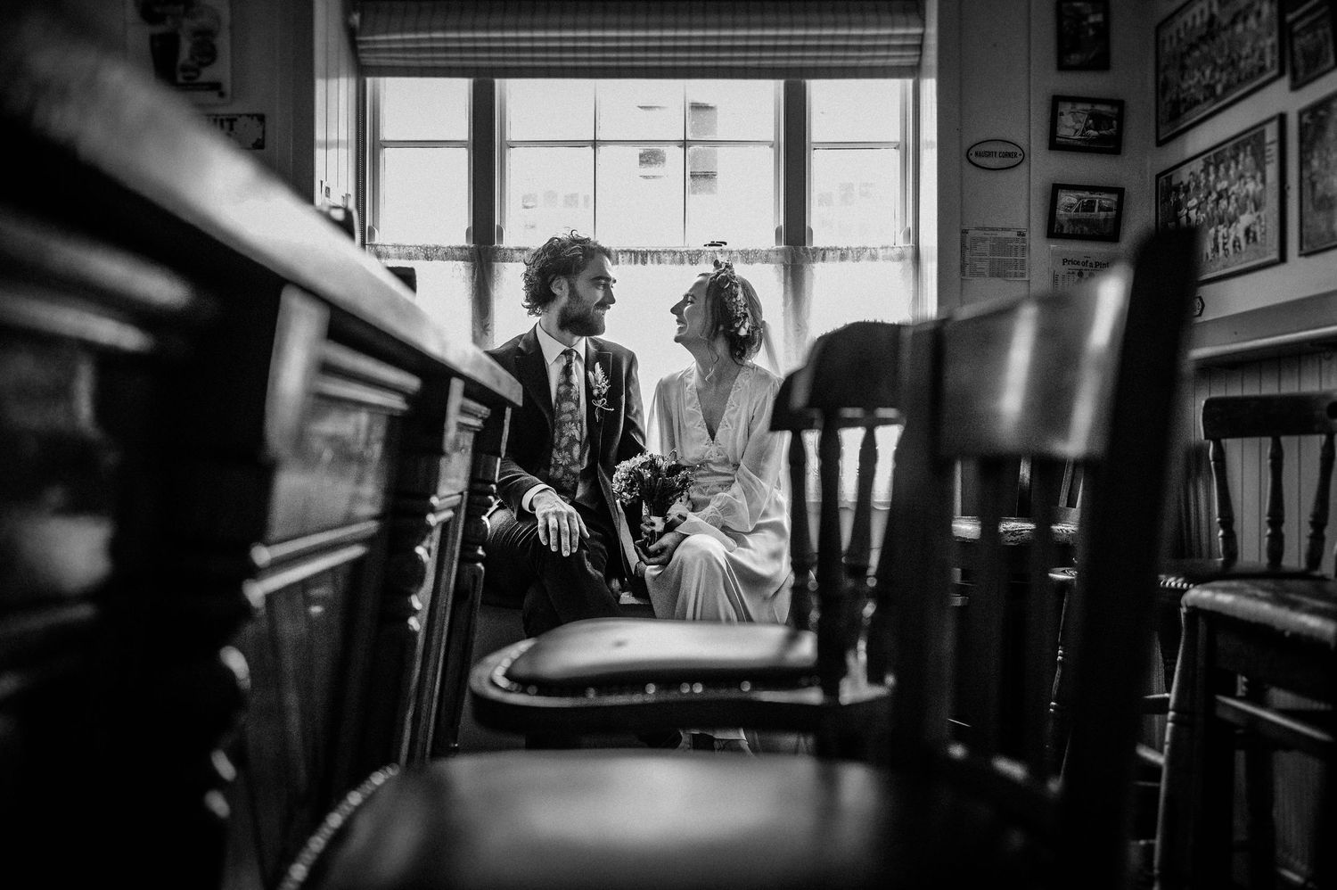 Black and white wedding photo of couple sharing a tender moment seated by a window in a rustic venue.