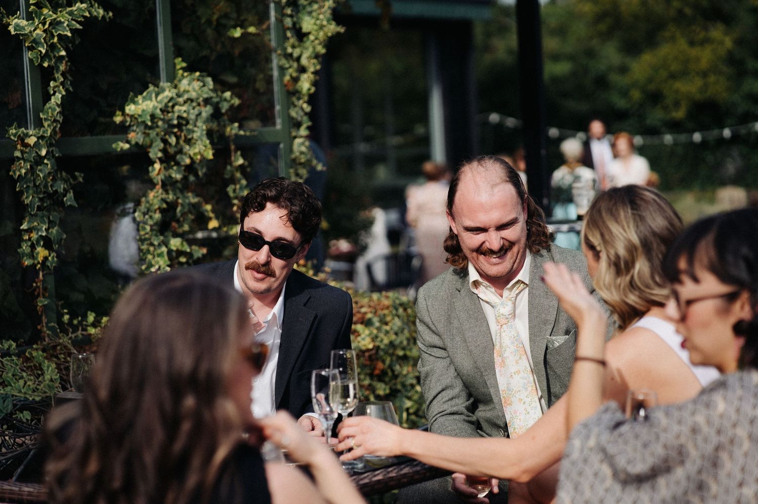 Guests laughing and toasting with champagne glasses at an outdoor wedding reception on a sunny day.