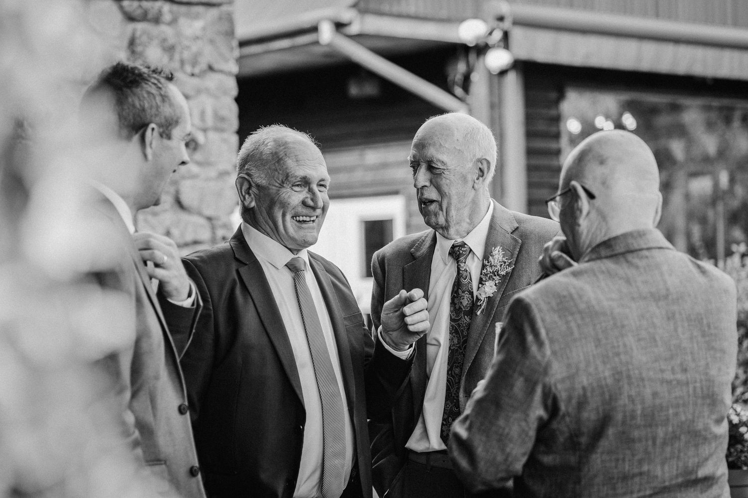 Black and white photo of elderly men in suits laughing and conversing at a wedding reception outdoor venue.