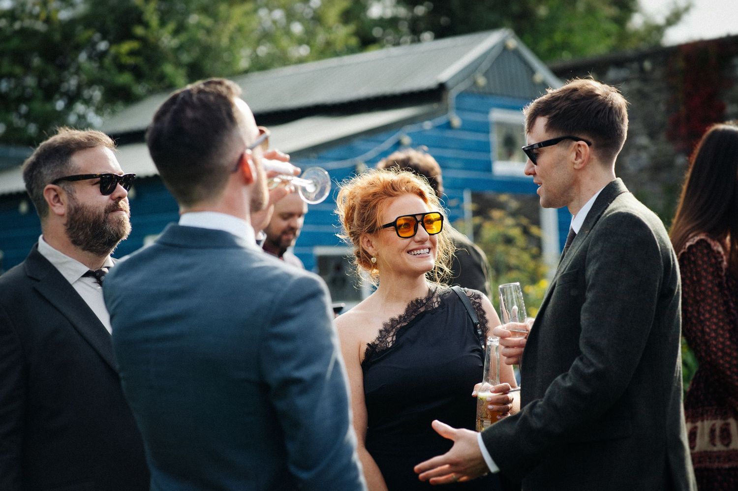 Elegantly dressed guests socializing outdoors at a wedding reception, woman in black dress wearing sunglasses.