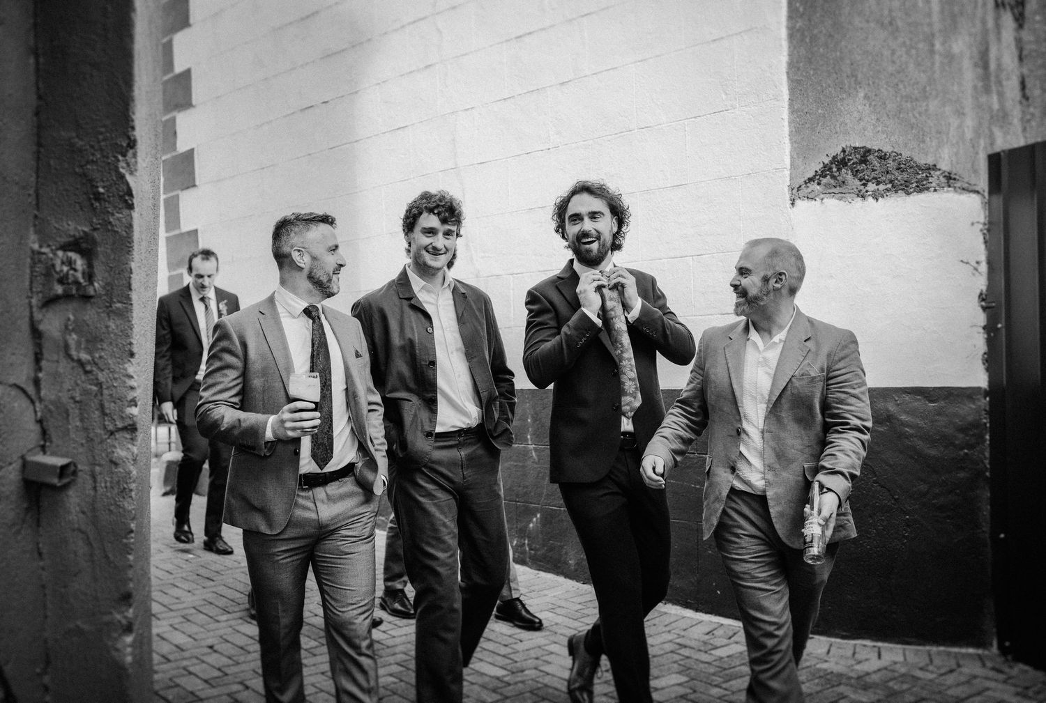 Black and white photo of four men in suits laughing and walking together outdoors.