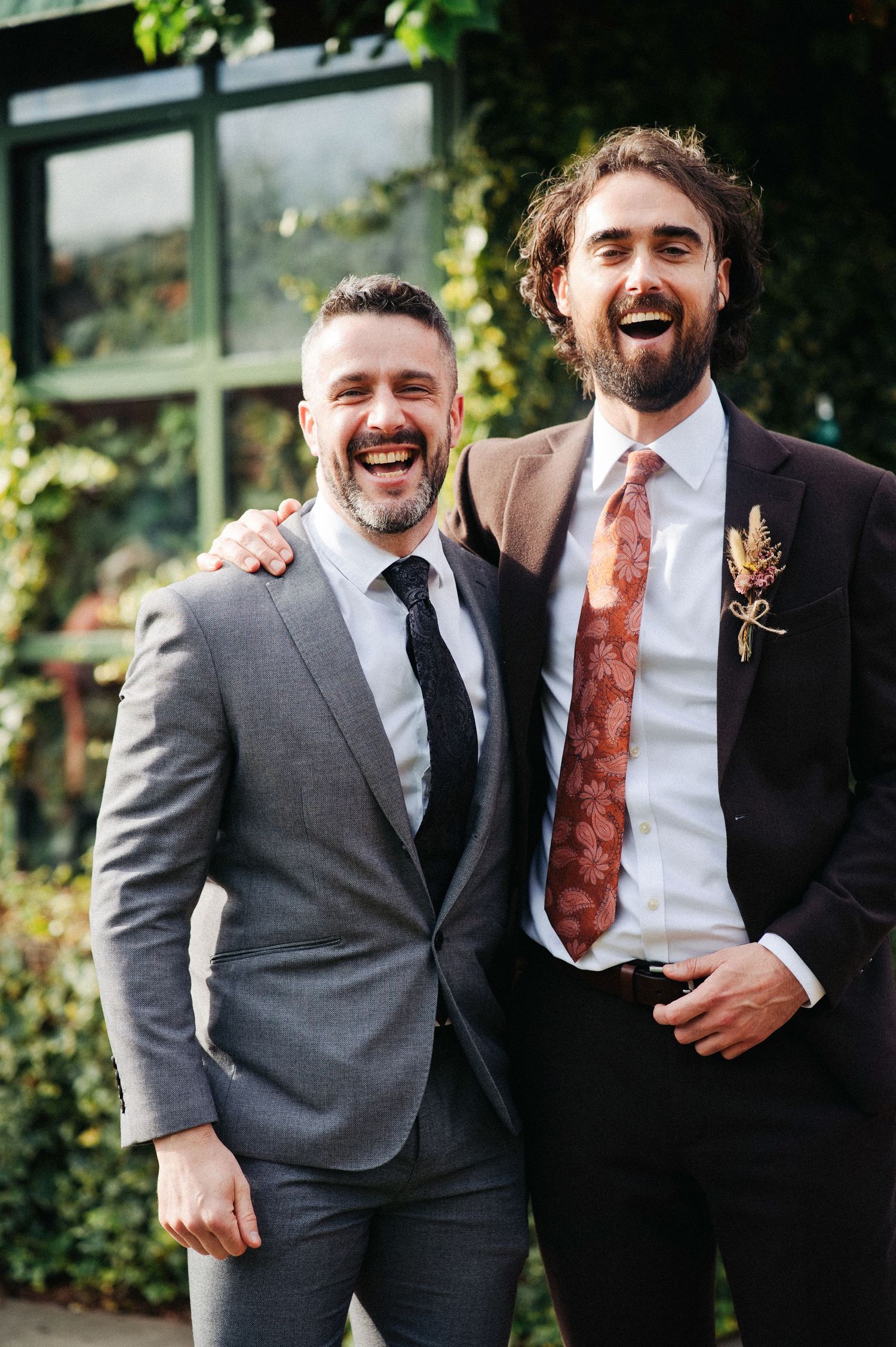Two smiling grooms in suits with ties laugh together outdoors at their wedding celebration.