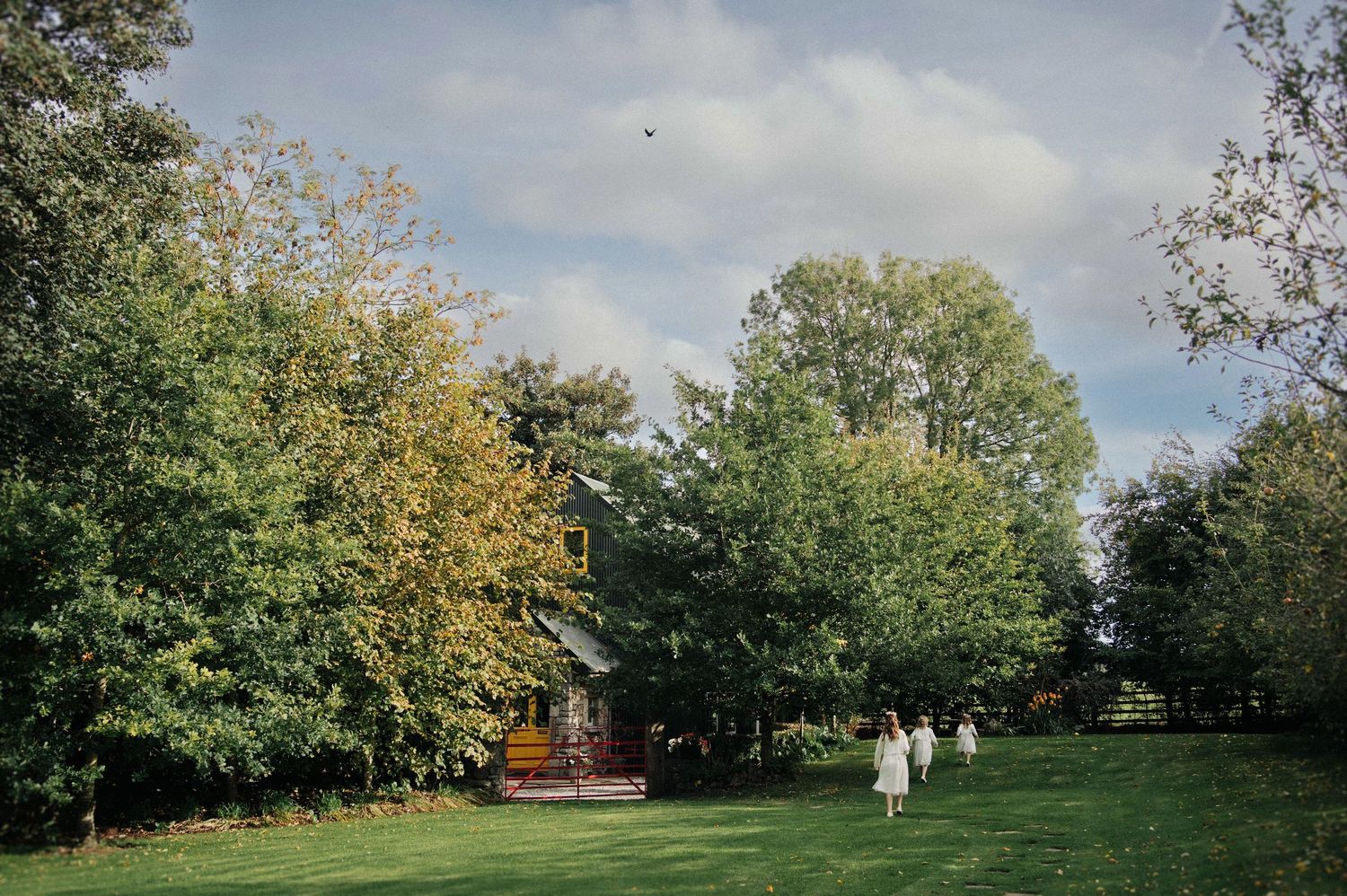 Lush green garden with tall trees surrounding a charming countryside barn venue on a partly cloudy day.