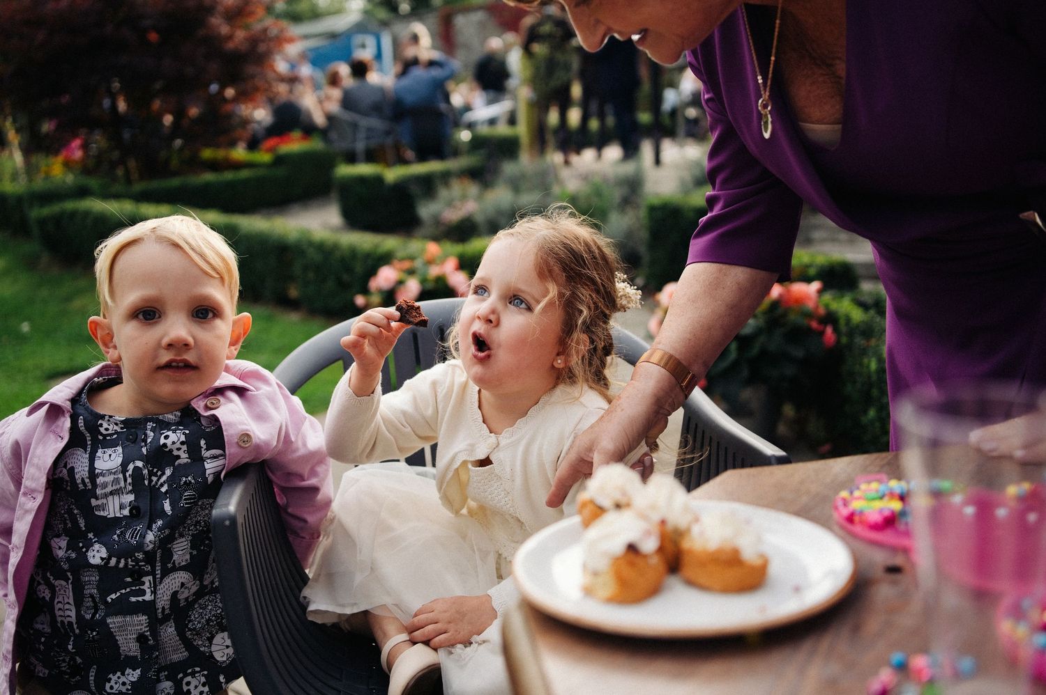 Two young children sitting outdoors at a party table with cupcakes, as an adult offers them a treat.