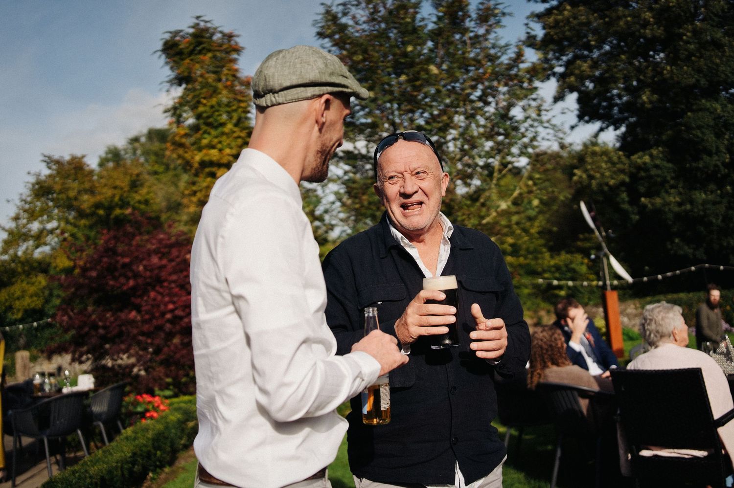 Two men laughing and holding drinks at an outdoor garden party on a sunny day.