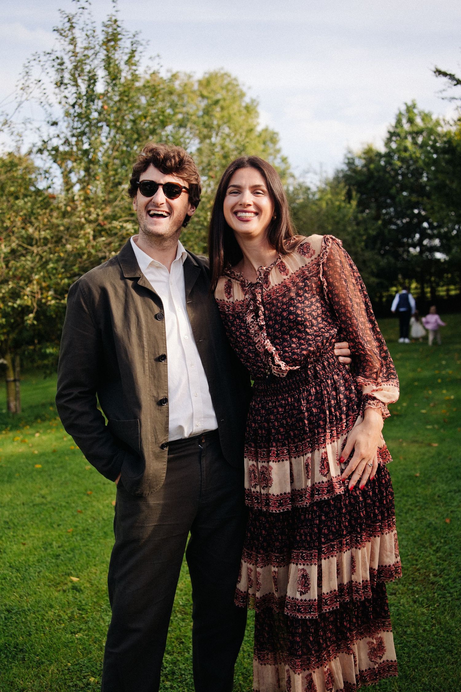 Smiling couple posing outdoors in a lush green garden, man in dark jacket with sunglasses, woman in floral maxi dress.