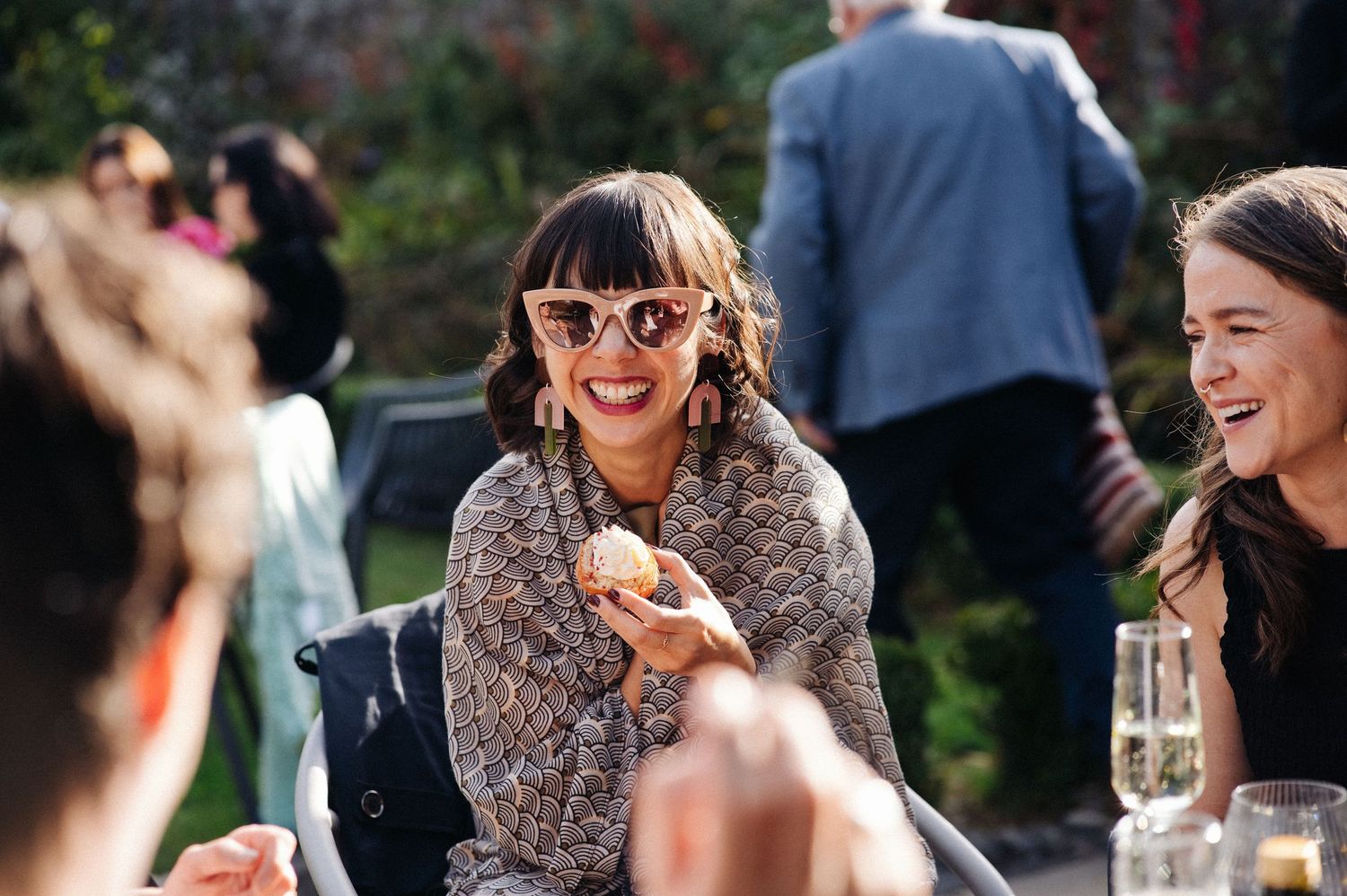 Women socializing outdoors at a sunny garden party, smiling and sharing food with champagne glasses on the table.