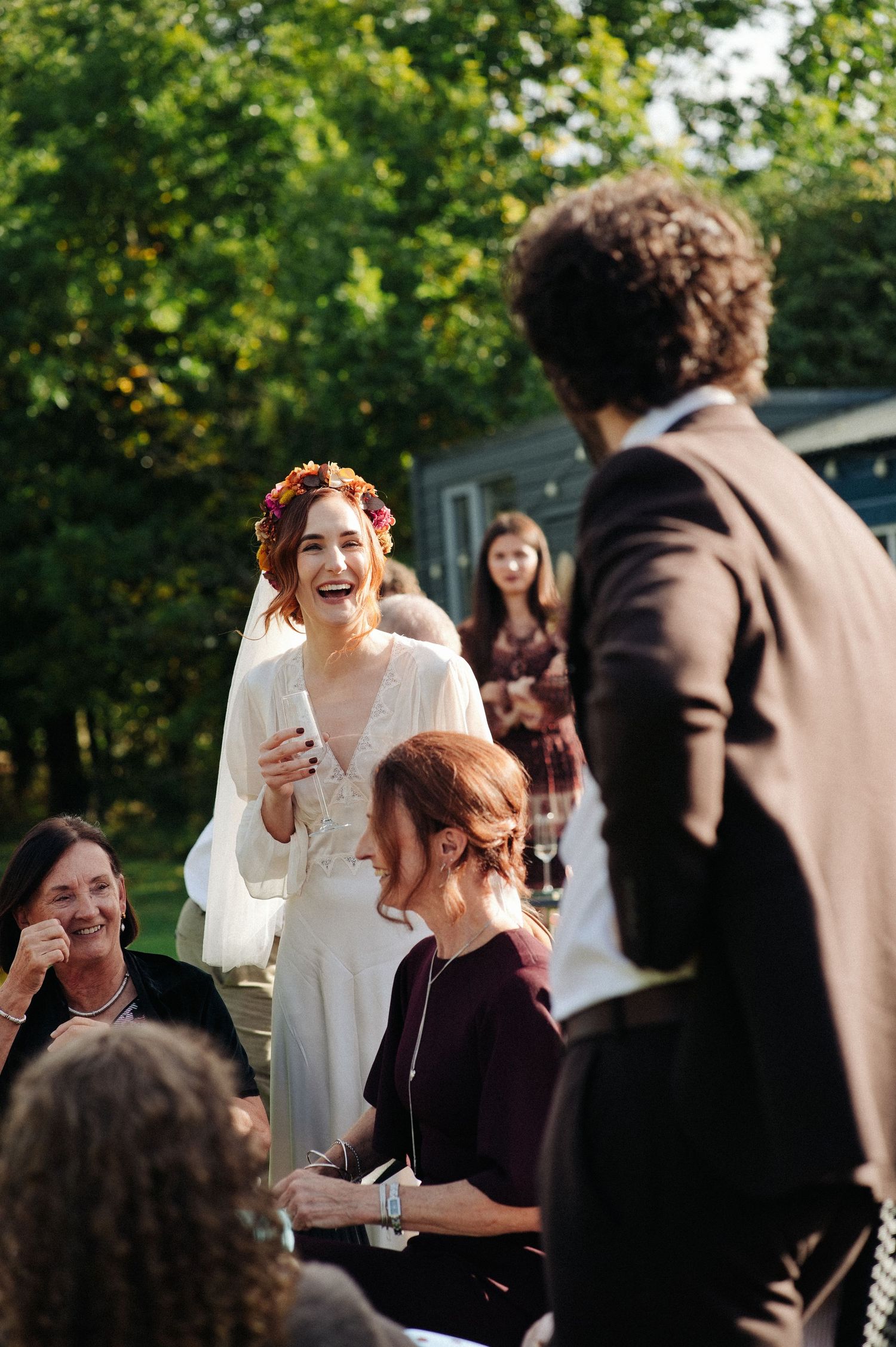 Bride in white dress and floral crown laughing joyfully with guests at an outdoor wedding reception.