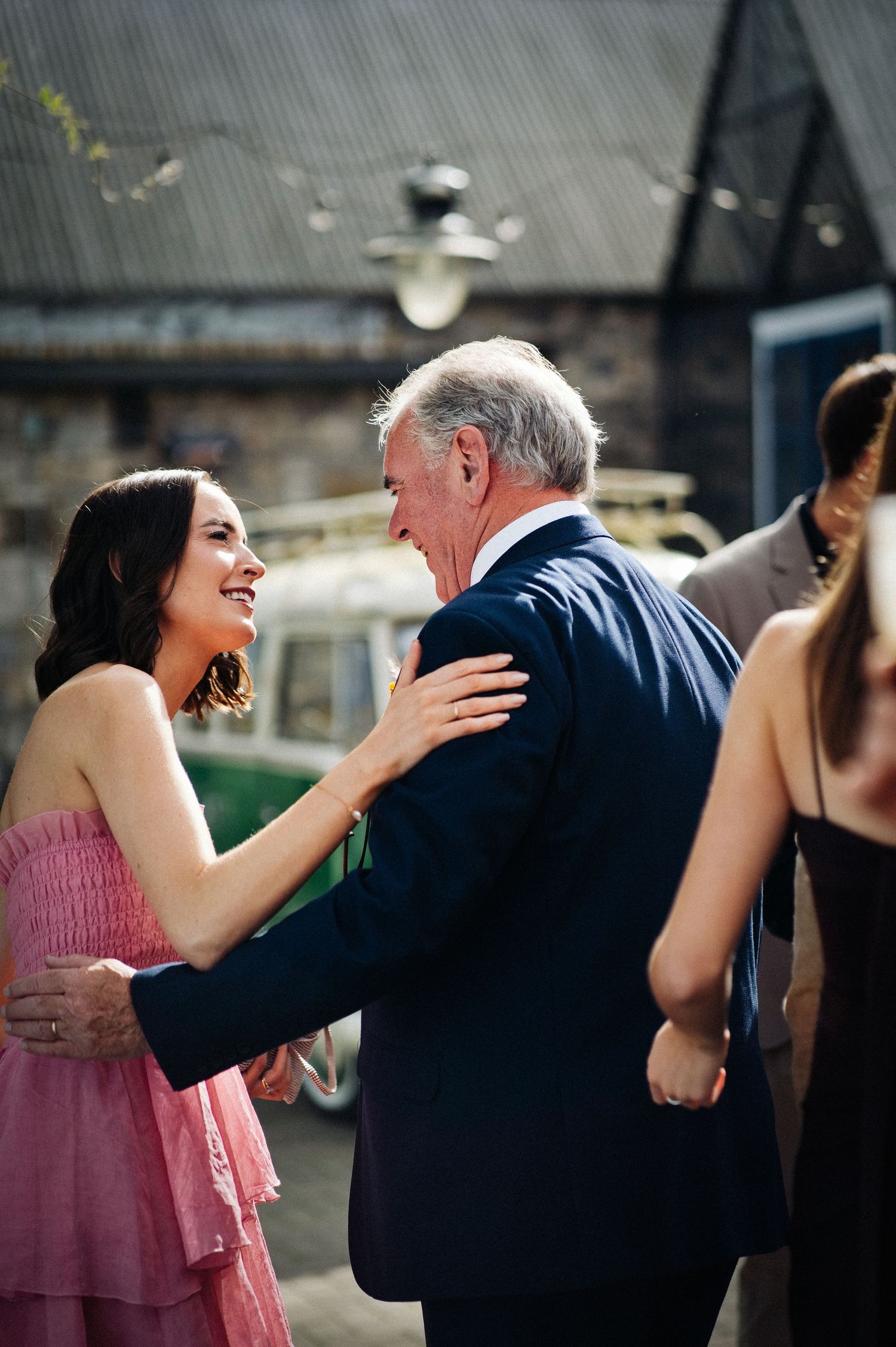 A young woman in a pink dress smiles warmly while embracing an older man in a navy suit at an outdoor event.