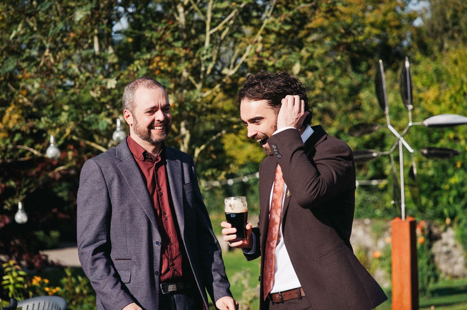 Two men in suits socializing outdoors at a garden event, one holding a dark beer glass on a sunny day.