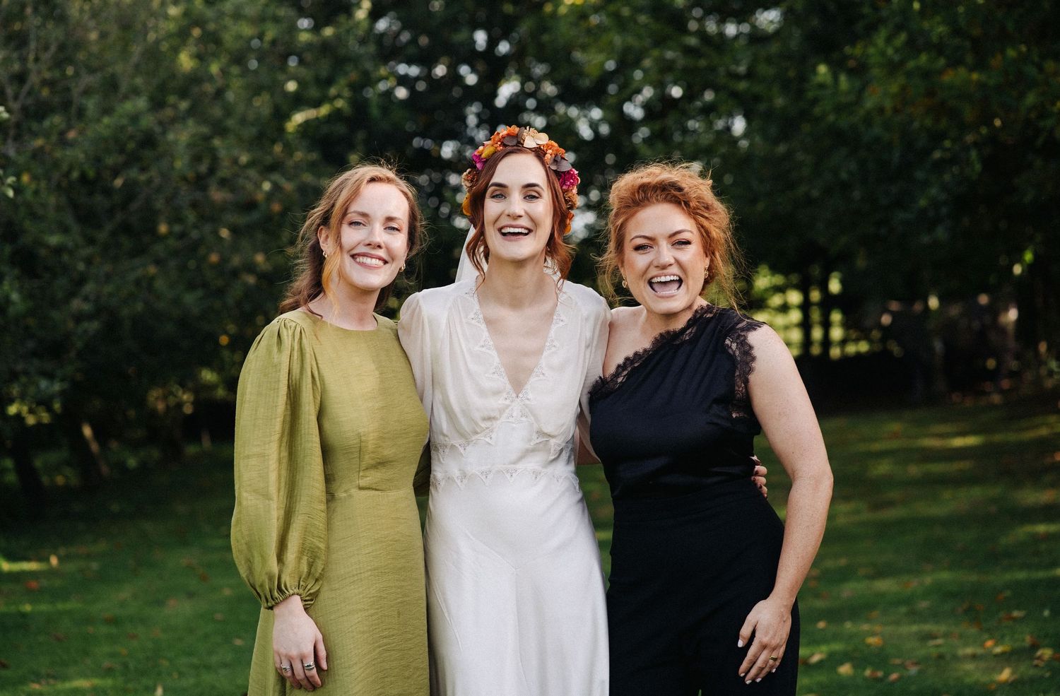 Three women smiling outdoors at a wedding, bride in white with floral crown flanked by guests in green and black dresses.