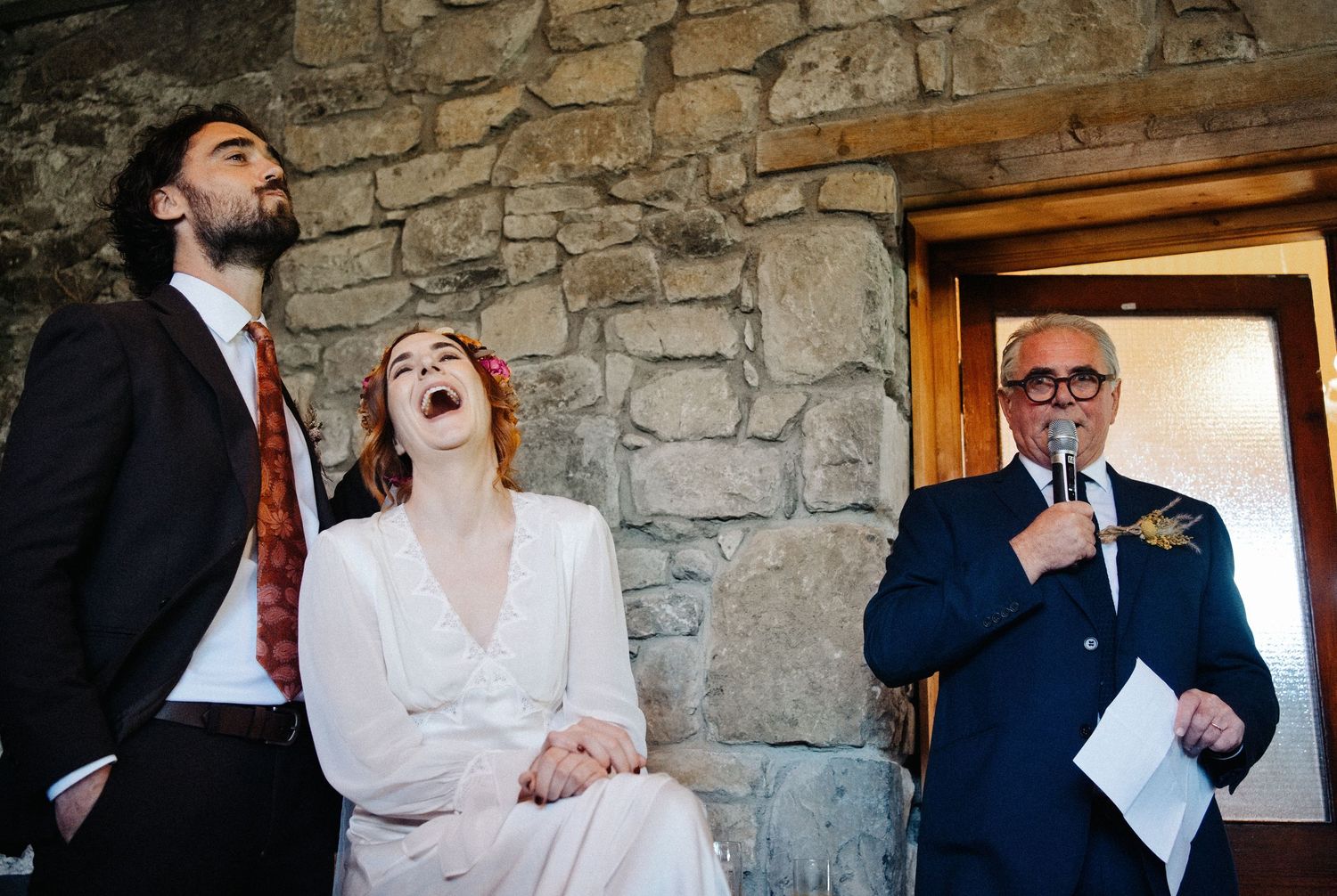 Bride laughing joyfully during wedding speech while groom and elderly man with microphone look on, stone wall backdrop.