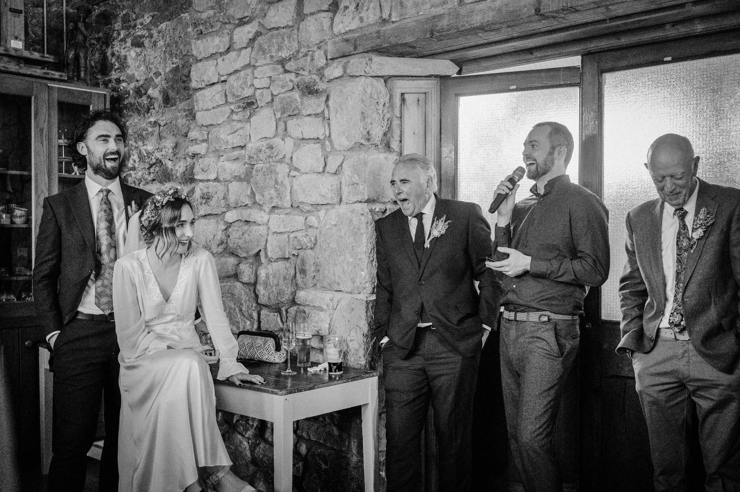 Black and white wedding reception photo with guests listening to a speech by a man holding a microphone indoors.
