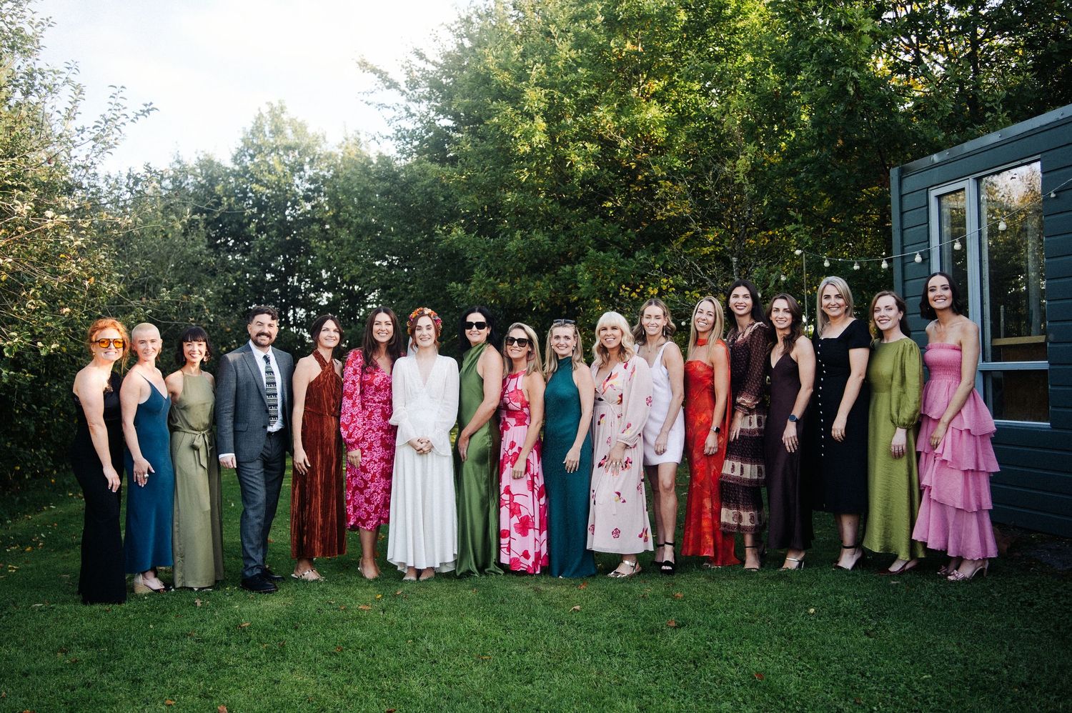 Large group of elegantly dressed wedding guests posing together on green lawn surrounded by lush trees.