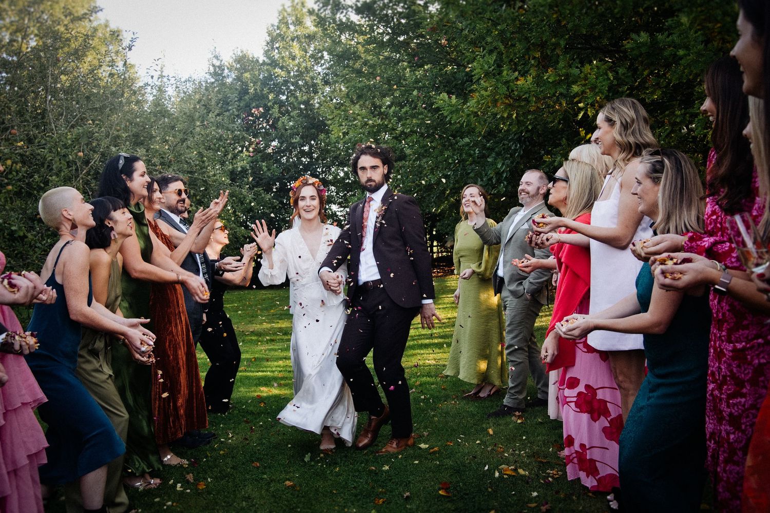 Bride and groom walk through cheering guests outdoors at garden wedding ceremony with lush green trees.