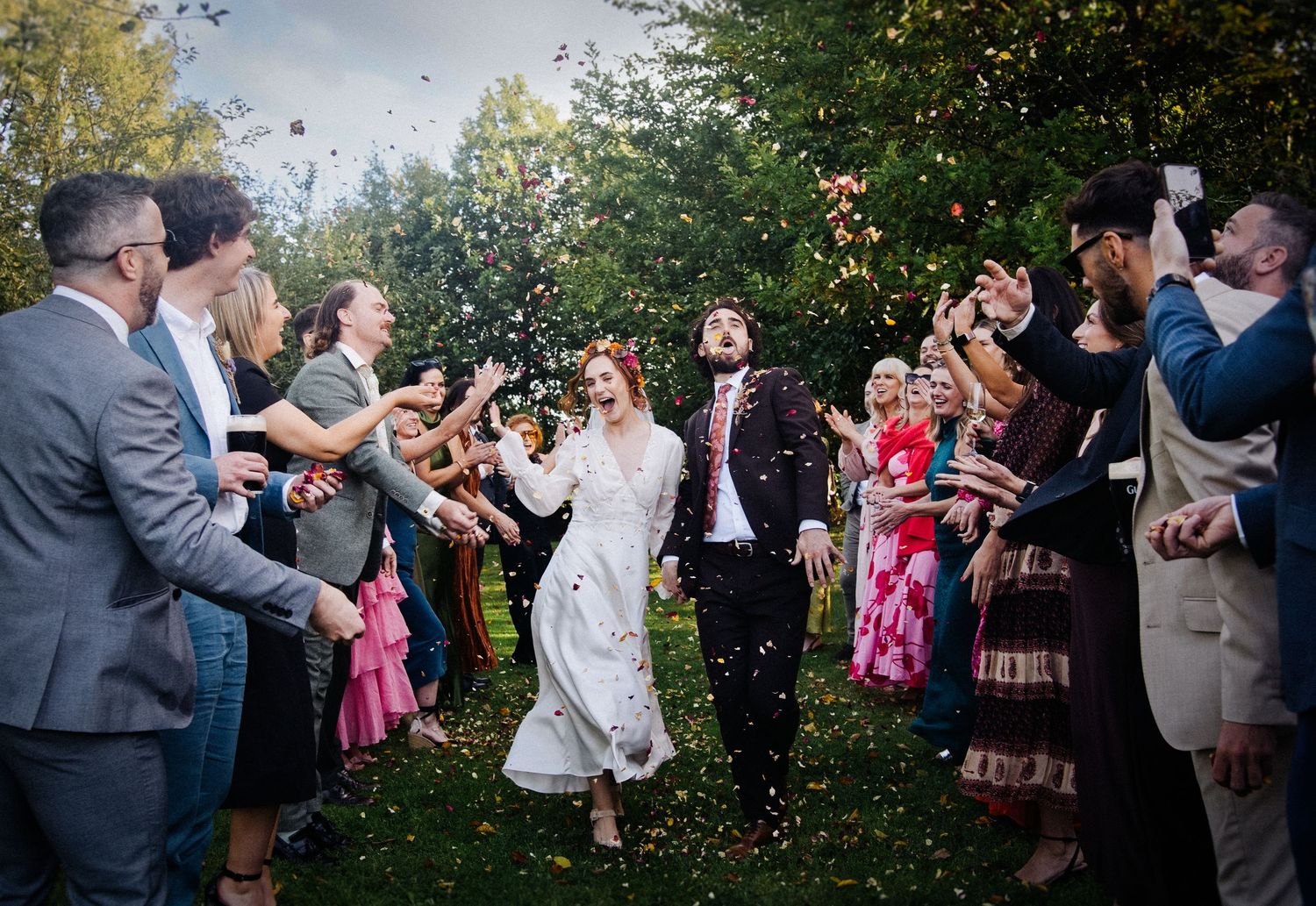 Joyful newlyweds walk through confetti shower surrounded by cheering wedding guests outdoors.