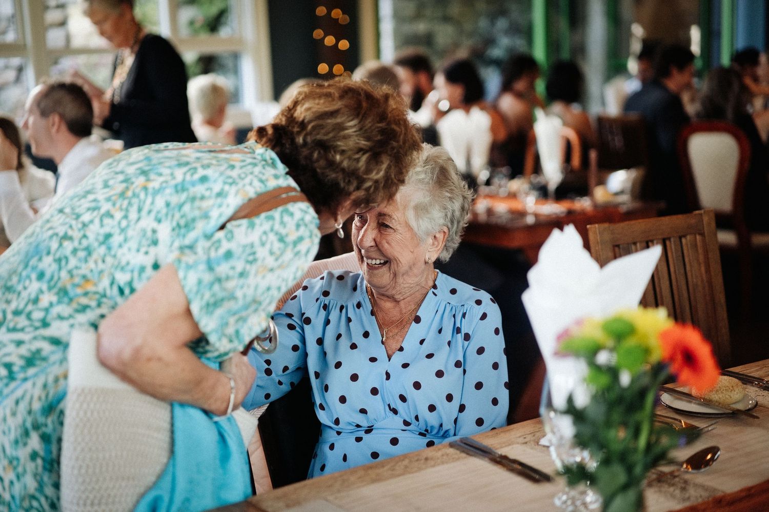 Two elderly women share a warm moment at a restaurant table, one leaning in affectionately while the other smiles joyfully.
