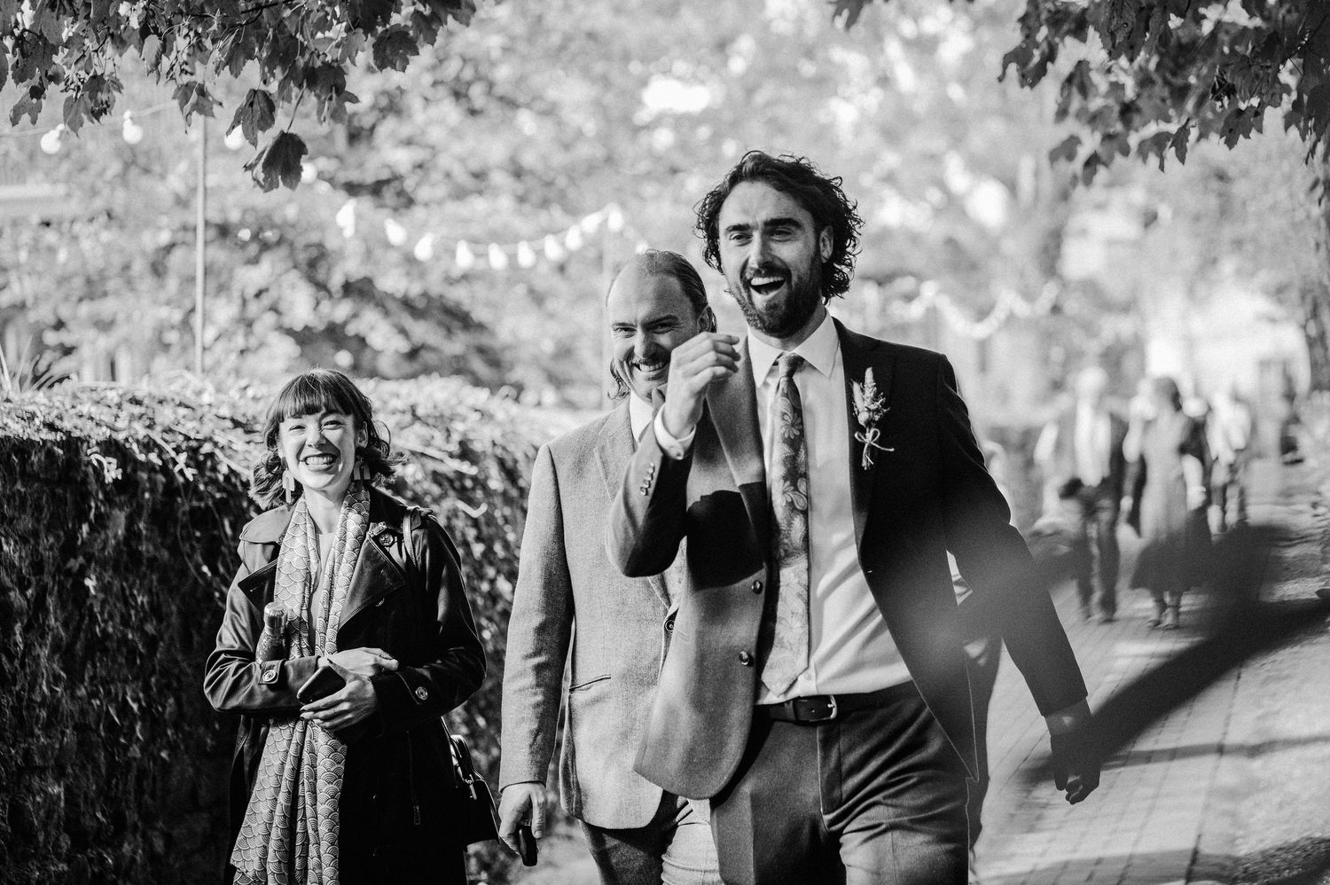 Black and white photo of smiling groom walking with guests at outdoor wedding celebration.