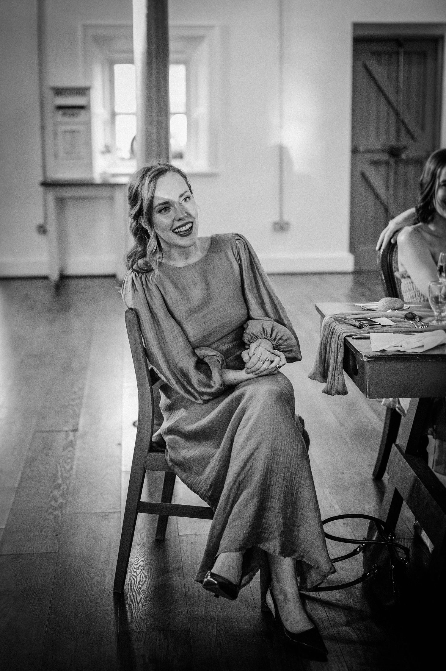 A smiling woman in a long gray dress sits on a wooden chair at an indoor event in a black and white photo.