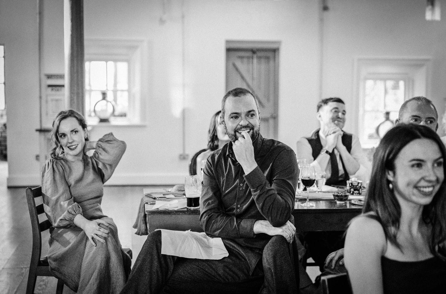 Black and white photo of wedding guests laughing and reacting during speeches at an elegant indoor reception.