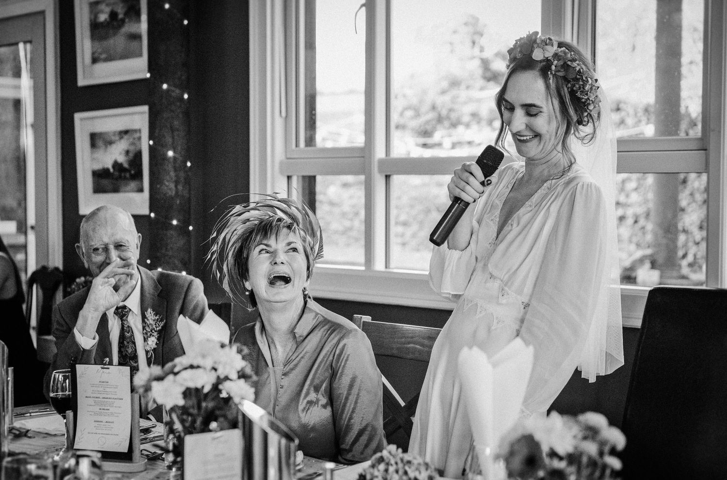 Bride gives a speech at wedding reception as guests laugh joyfully at a decorated table in black and white photo.