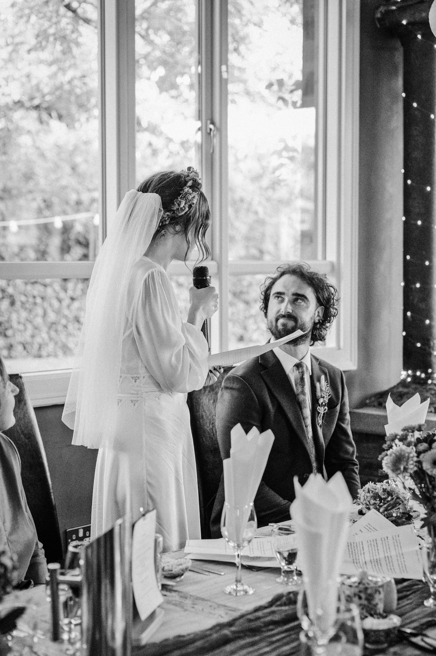 Bride giving a speech at wedding reception while groom sits at table, elegant black and white photography.