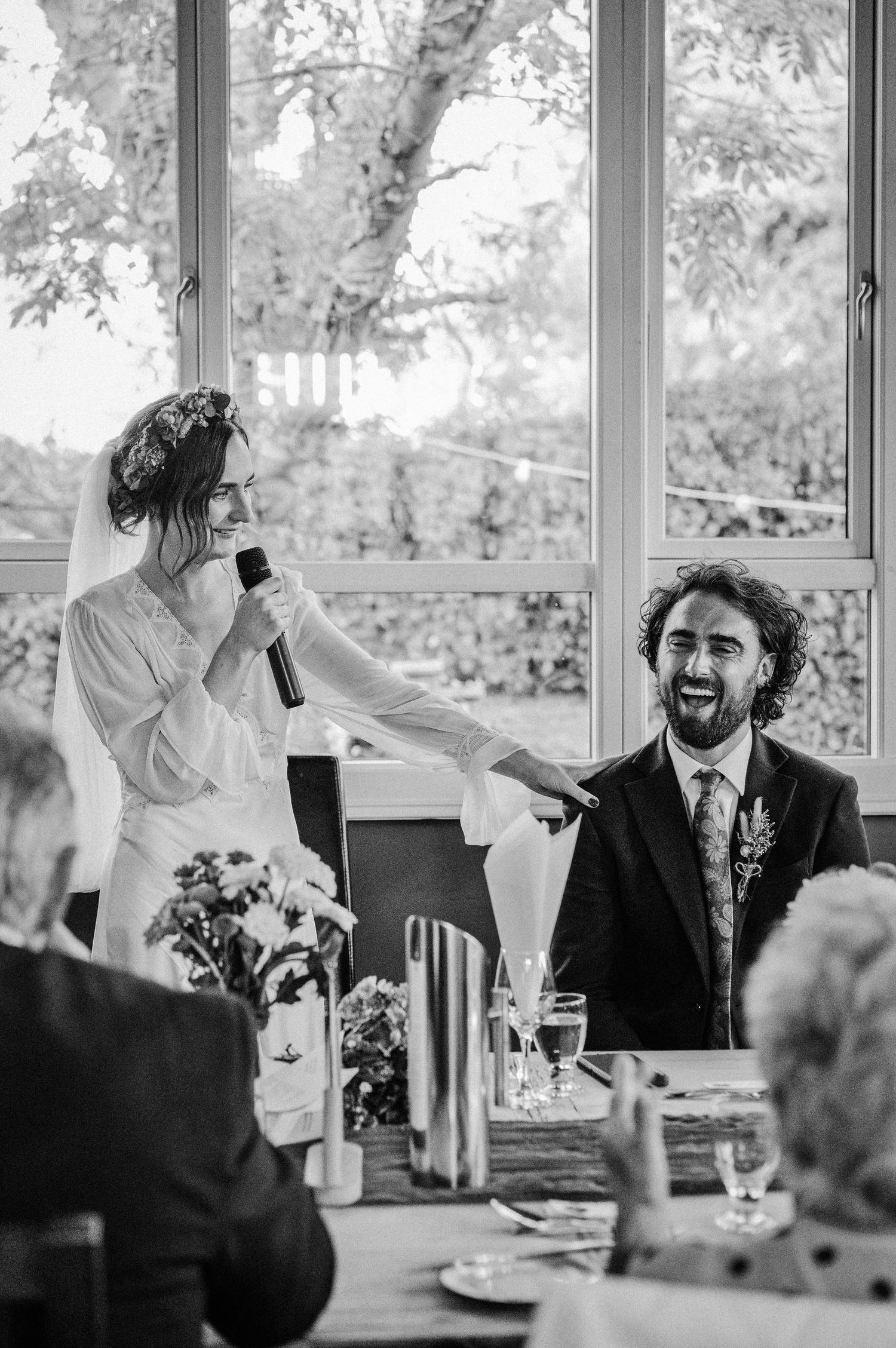 Bride holds microphone toward laughing groom during wedding reception speech at elegant venue with large windows.