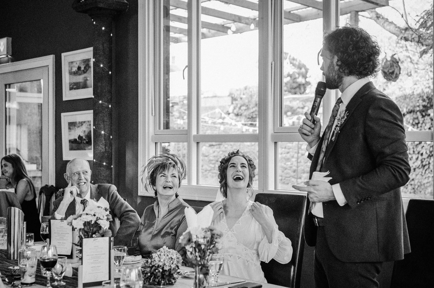 Black and white wedding reception photo of bride laughing joyfully as groom gives a heartfelt speech to guests.