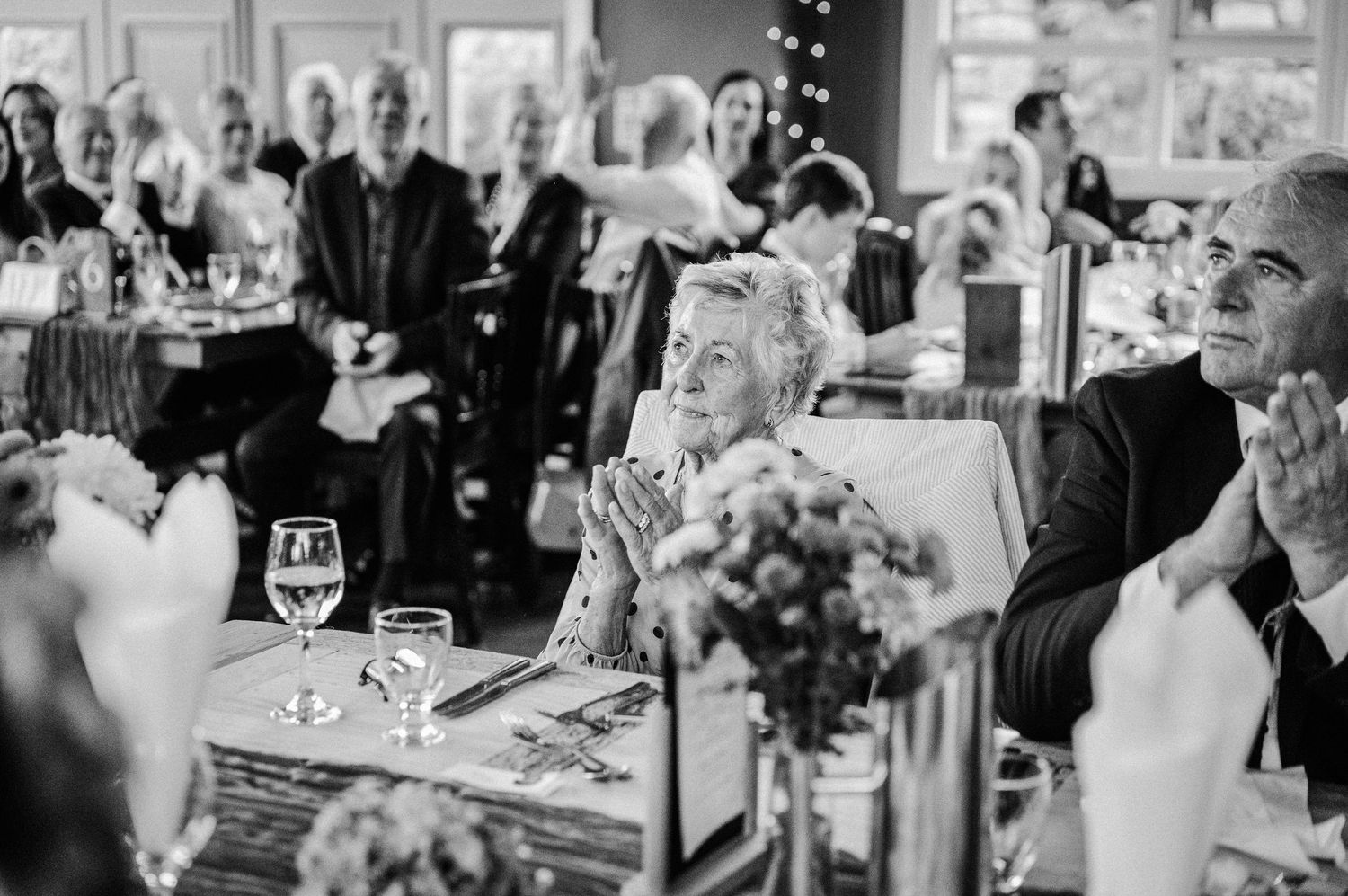 Elderly guests clapping and smiling at a wedding reception dinner, captured in black and white photography.