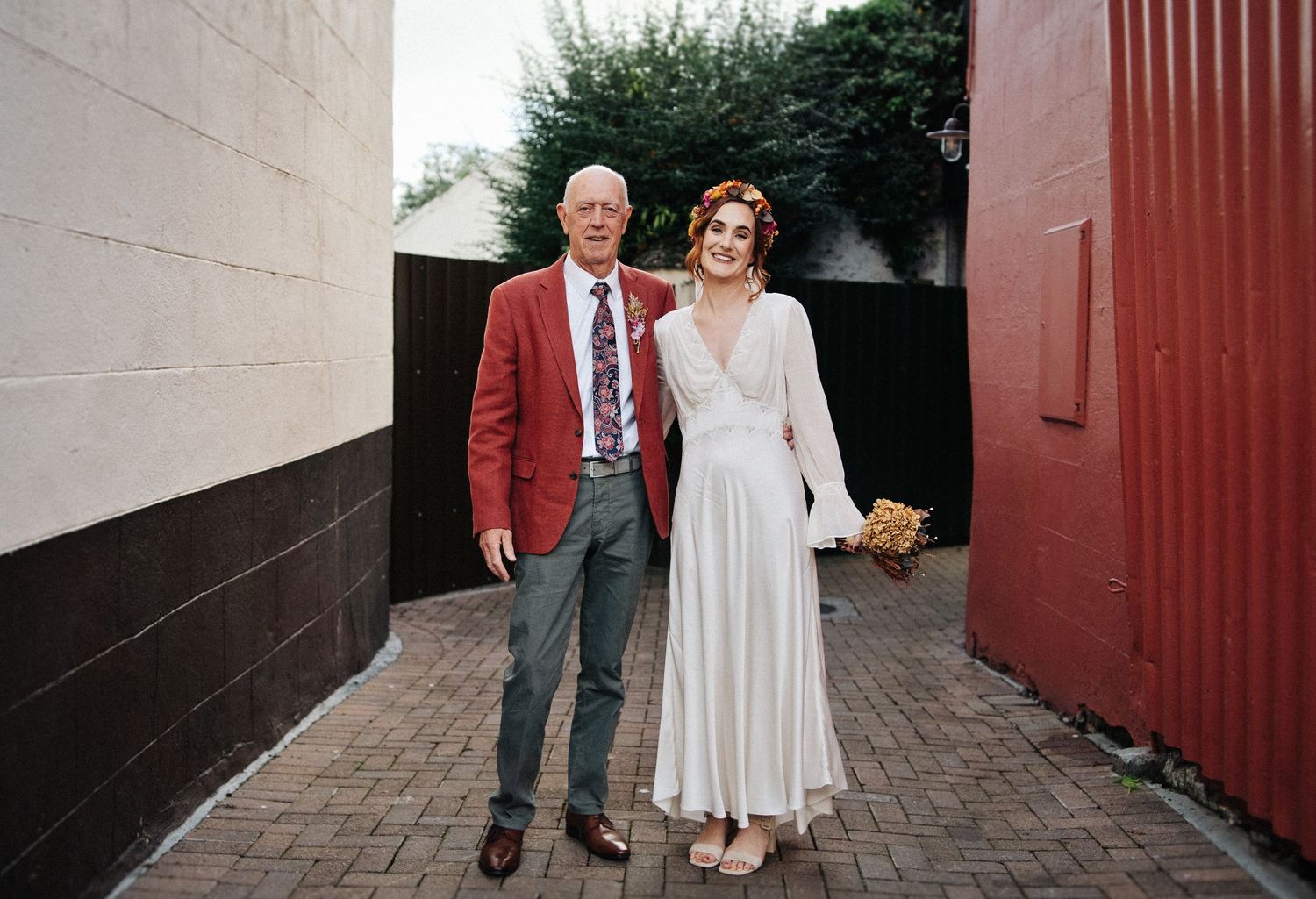 Bride in white dress and older man in red blazer posing together in a brick alleyway on a wedding day.