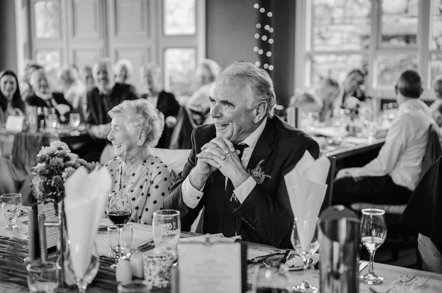 Black and white wedding reception photo of guests seated at tables, with an elegant man in a tuxedo smiling warmly.