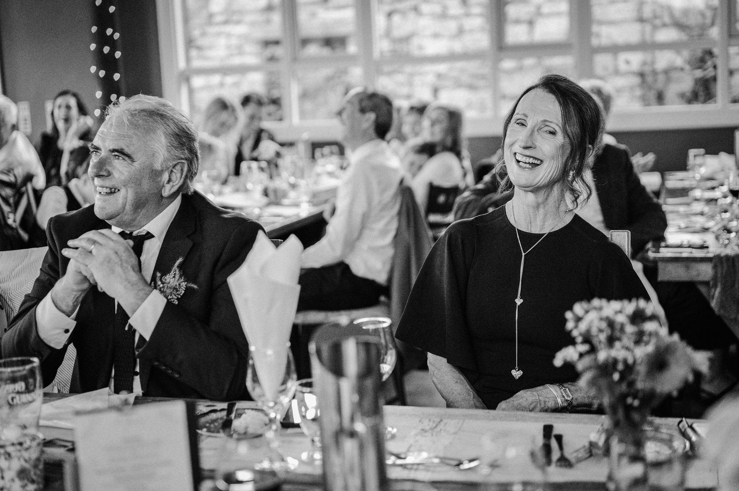 Guests laughing and smiling at a wedding reception dinner, captured in black and white photography.