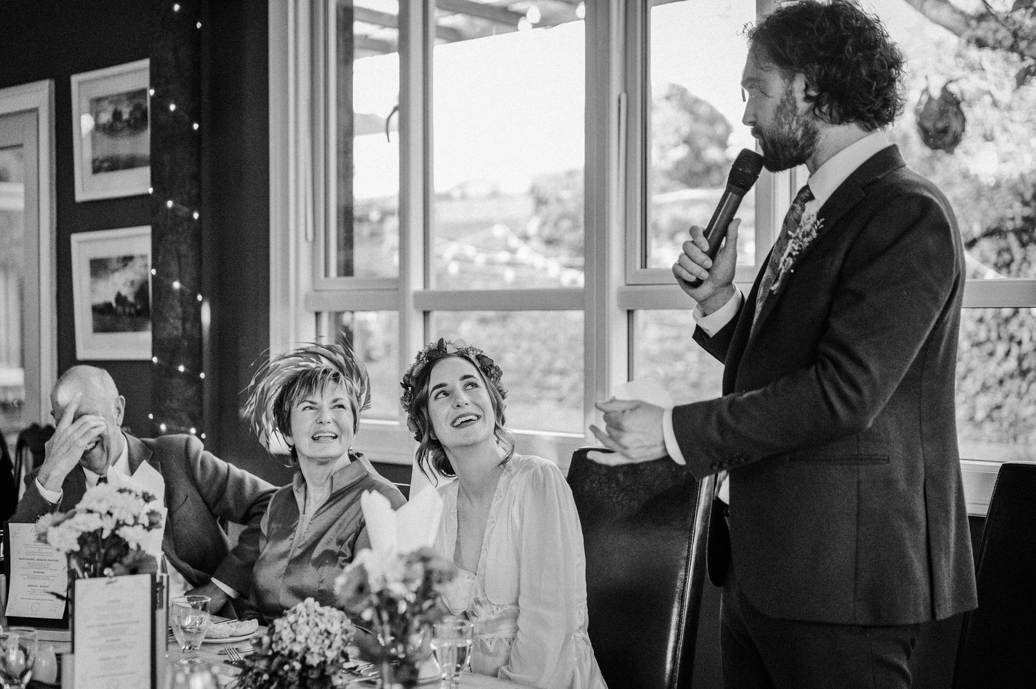 Black and white wedding reception photo of guests laughing during a heartfelt speech by a man in a suit.