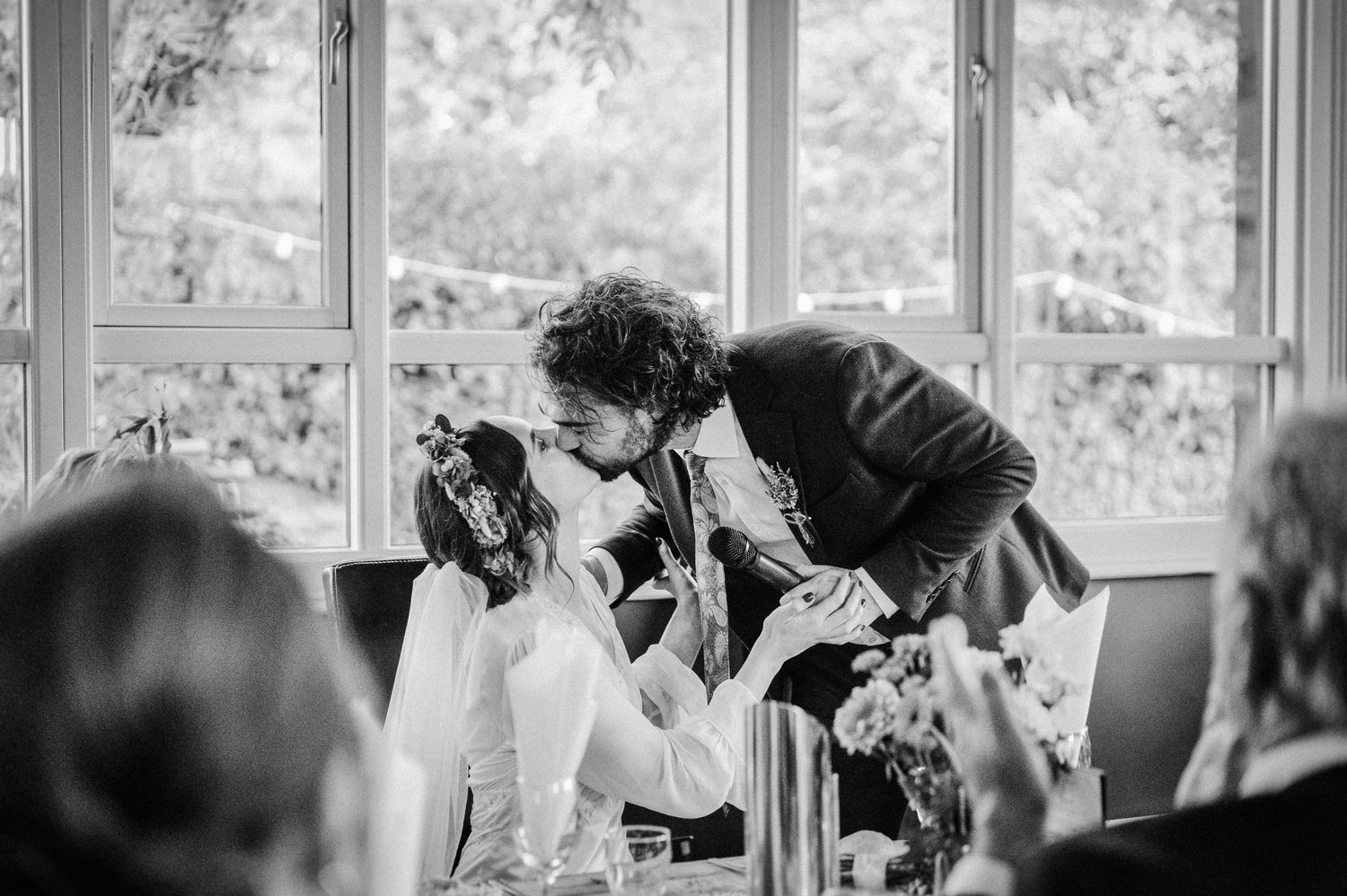 Black and white wedding photo of bride and groom sharing a kiss at the reception table near large windows.