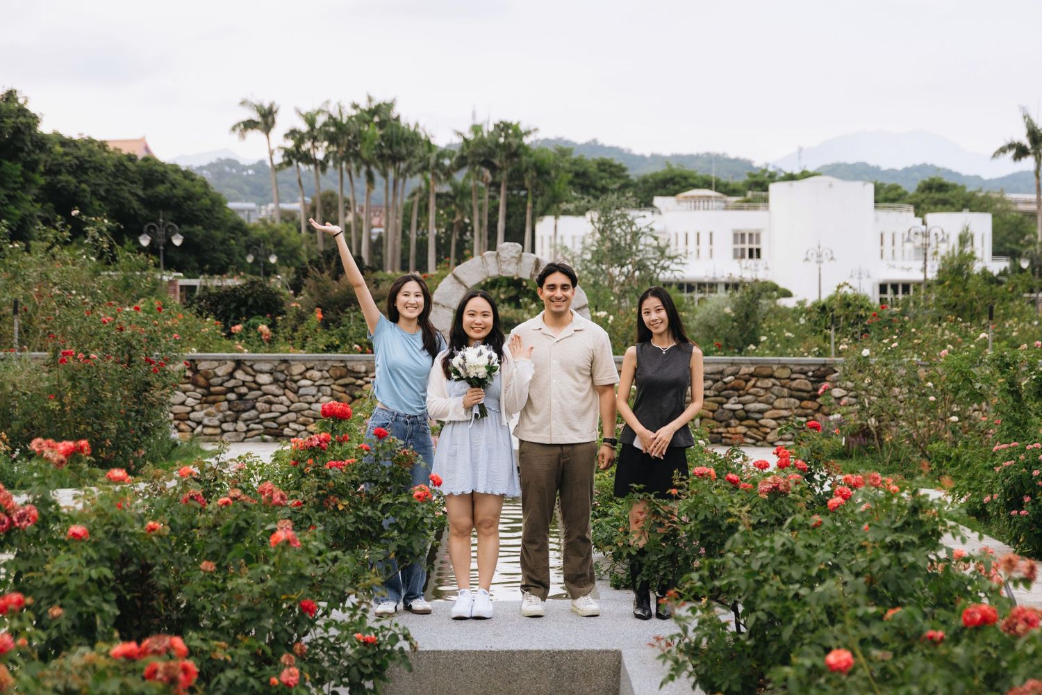 Four friends posing together in a beautiful rose garden with lush greenery and mountains in the background.