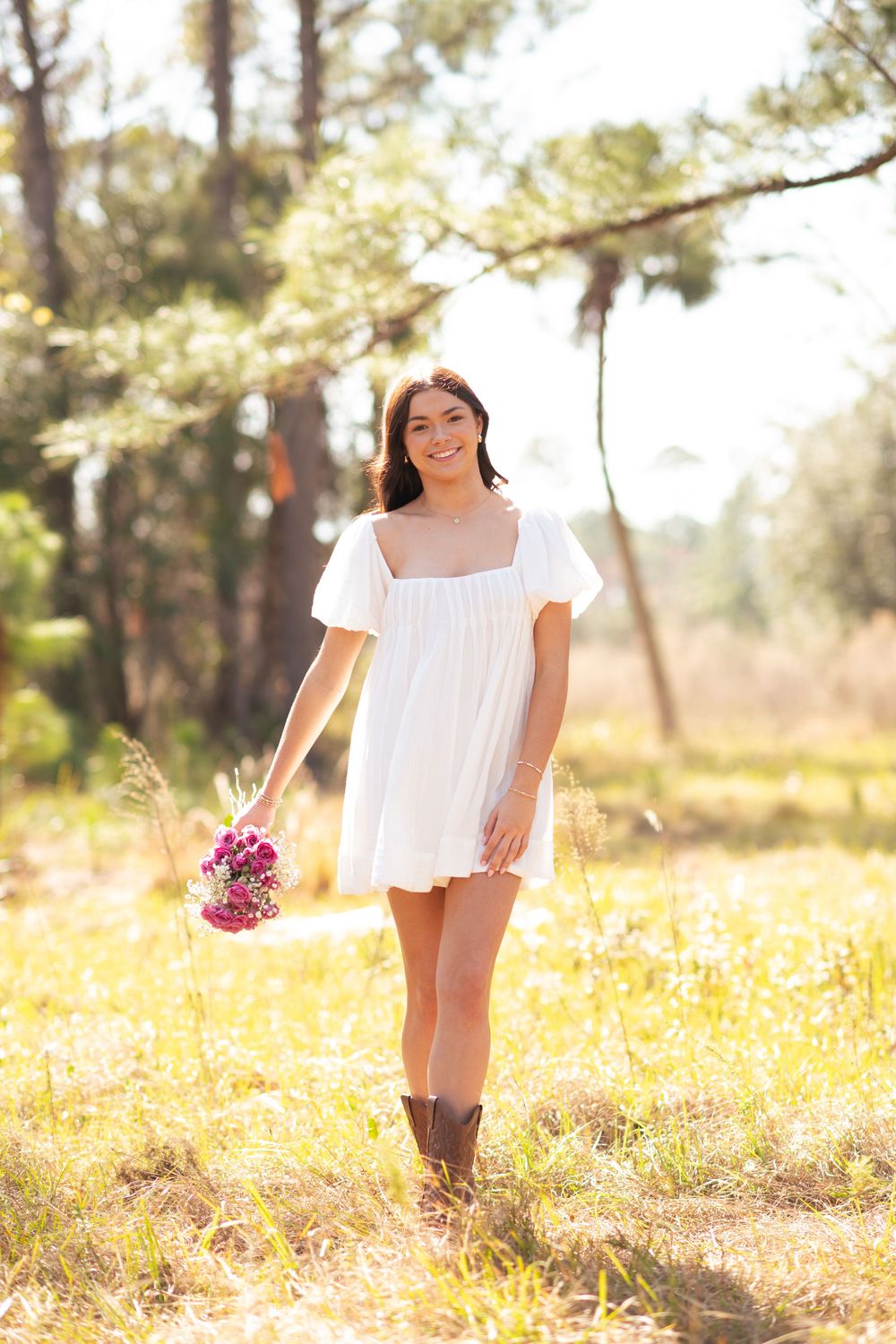 Young woman in white mini dress holding pink flowers, smiling in sunlit forest clearing wearing brown cowboy boots.