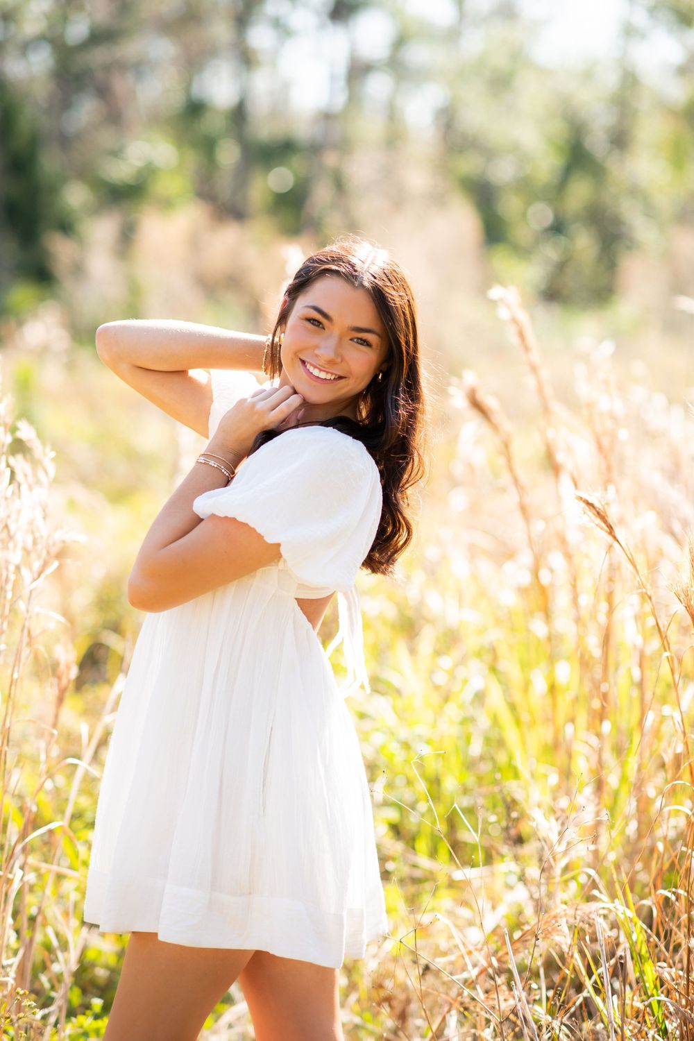 Young woman in white dress smiling in sunlit field with tall golden grasses and trees in background.