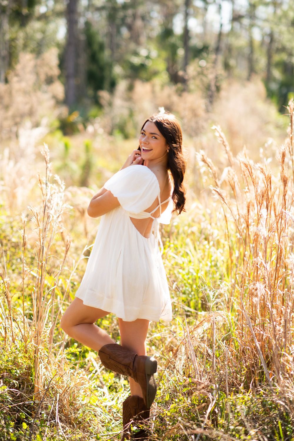 Young woman in white dress and brown cowboy boots smiling in a sunlit field of tall golden grasses.