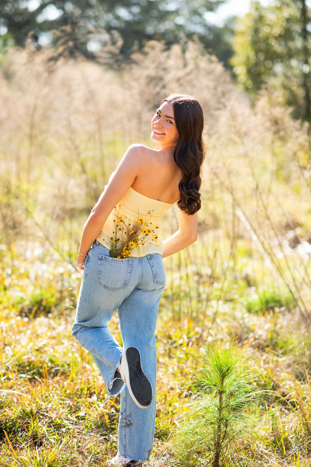 Young woman in yellow strapless top and jeans holds wildflowers, smiling outdoors in a sunlit field.